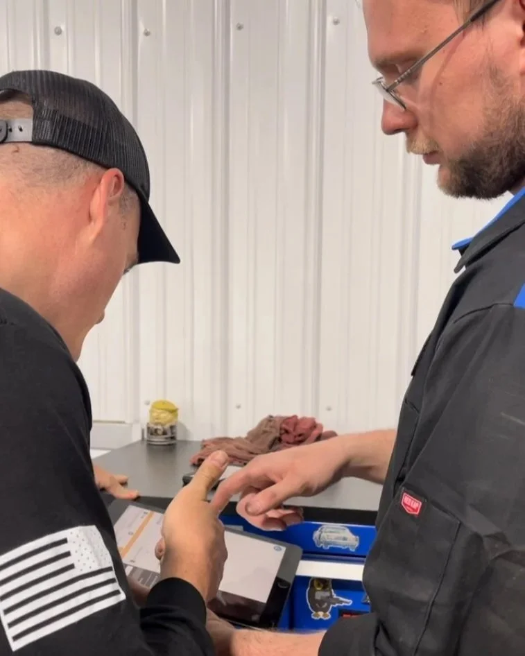 Two men looking at a tablet device, one holding it and the other pointing at the screen, in a workspace with a metal wall background, some folded clothes, and a jar on the table.