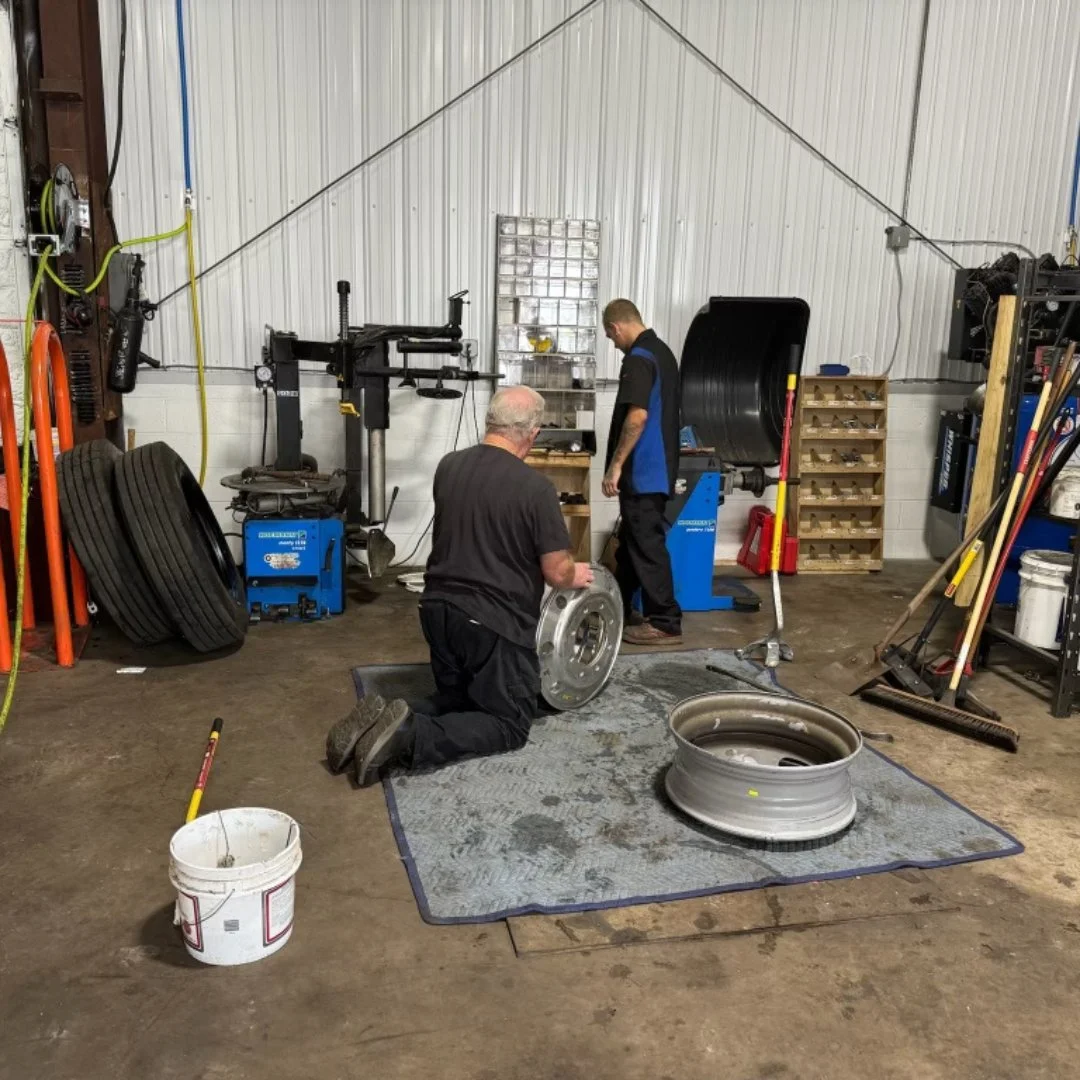 Two men working on a large metal wheel rim in a tire shop or wheel repair workshop. One man is kneeling and holding the rim, the other is standing and inspecting nearby. The workshop has tires, tools, and equipment organized on shelves and the floor.