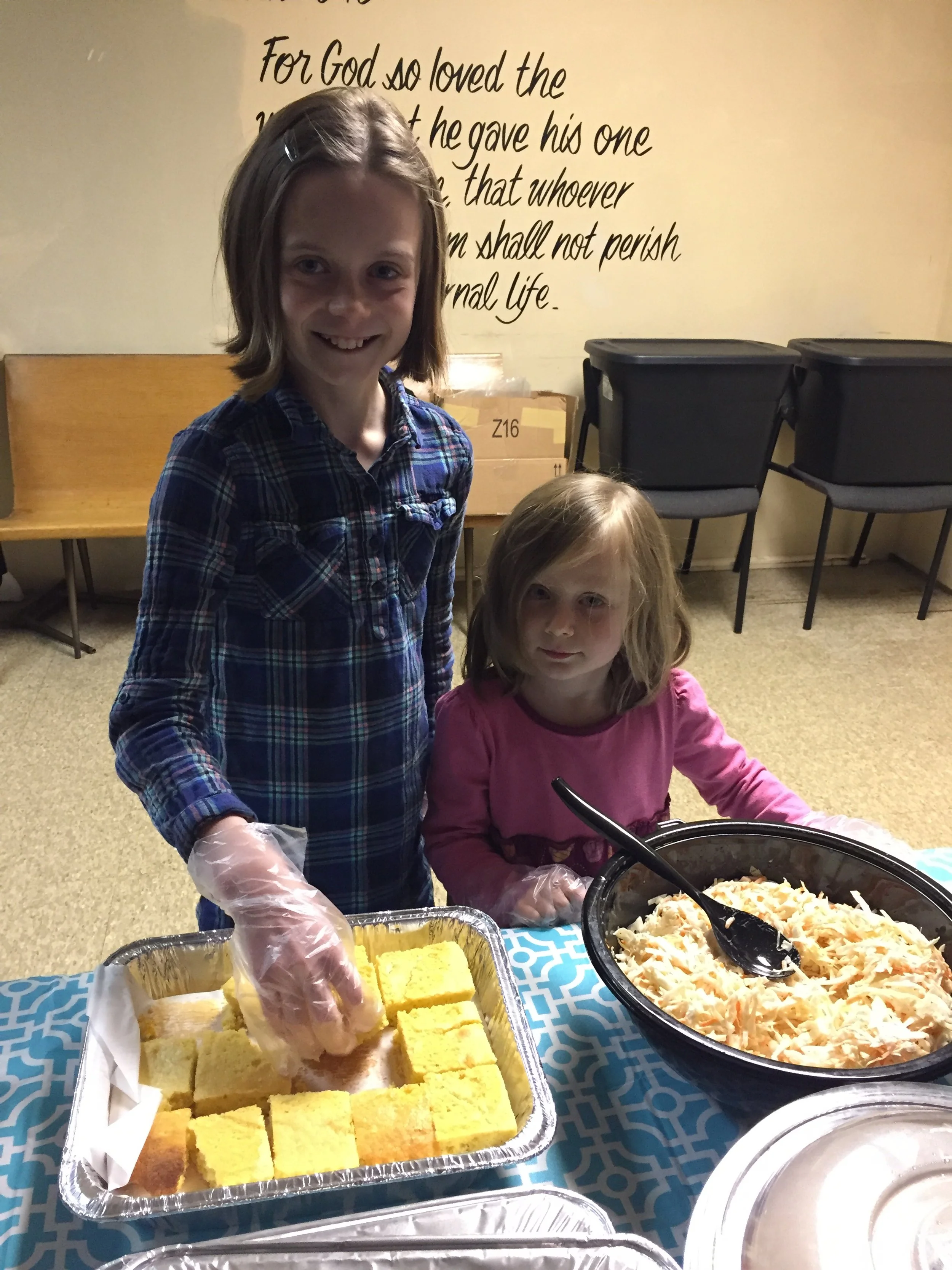 Two young girls are serving food at a table. The older girl is wearing a blue plaid shirt and putting pieces of cornbread from a foil tray onto a plate. The younger girl, wearing a pink shirt, is sitting next to large bowls of coleslaw. They are in a