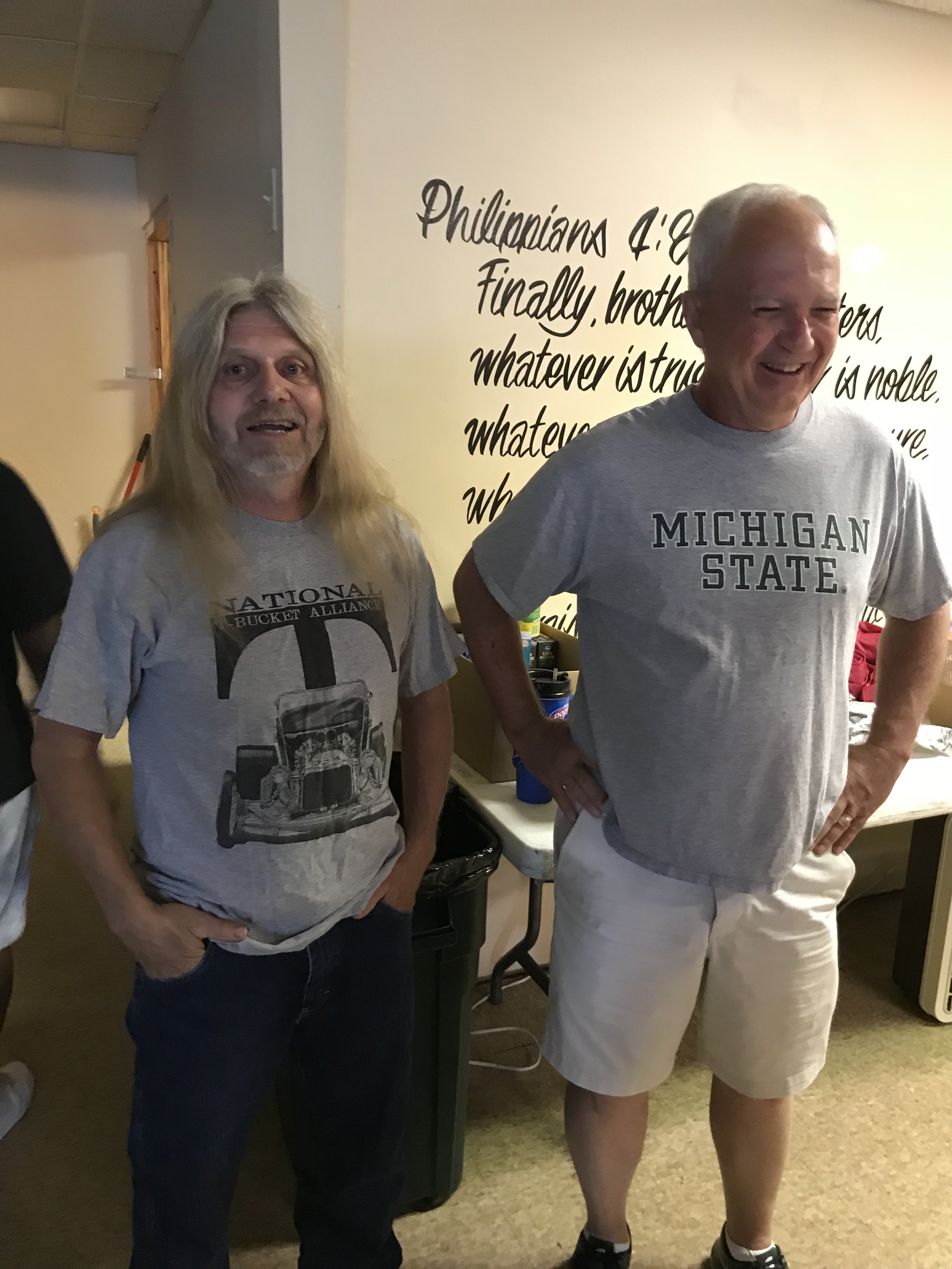 Two men standing and smiling indoors, one wearing a Michigan State T-shirt, the other wearing a gray shirt with a graphic. Background features a wall with a quote from Philippians 4:8.