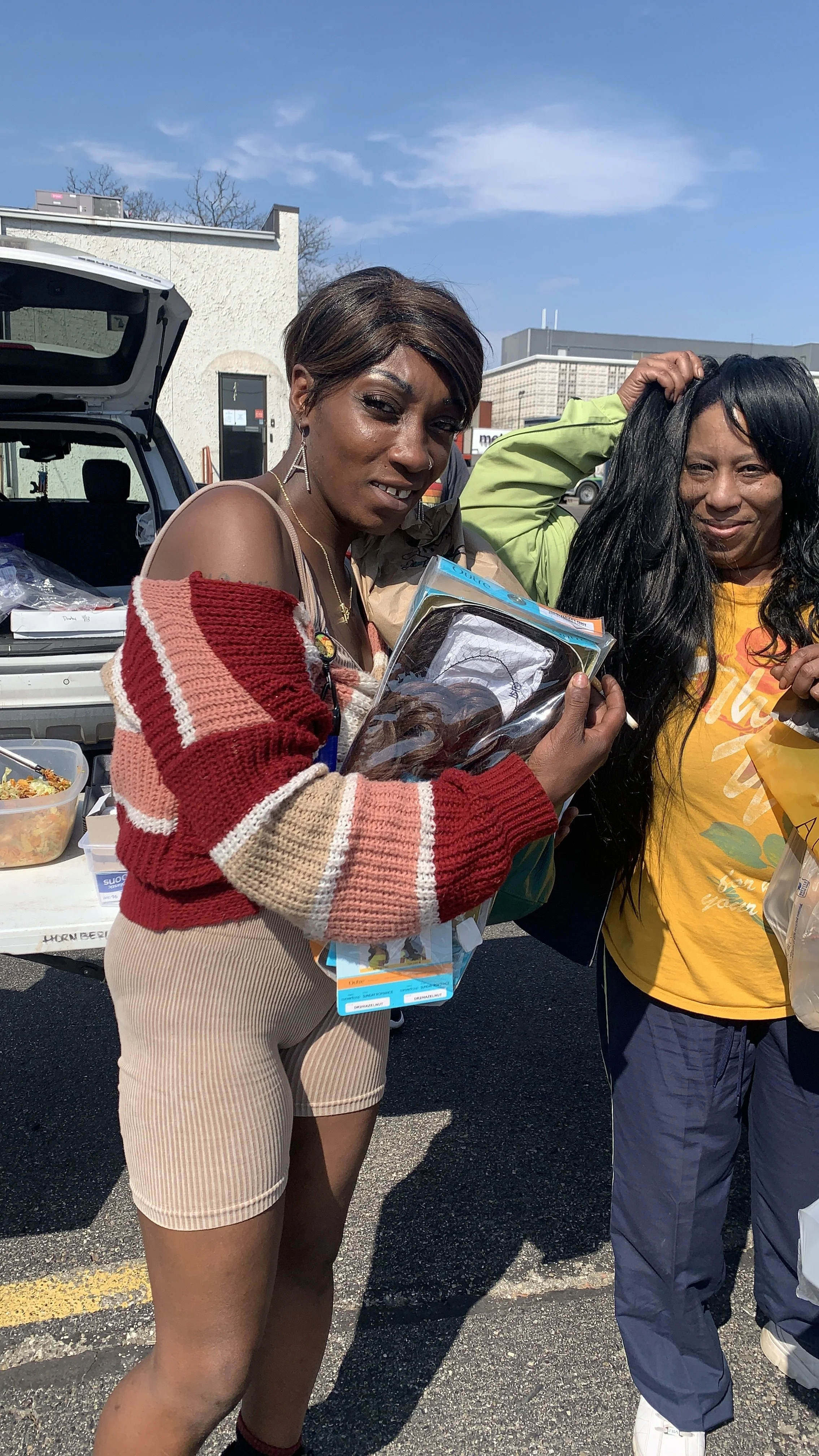 Two women standing outside near a car trunk filled with food items, one woman holding a Care Bears toy in plastic packaging.