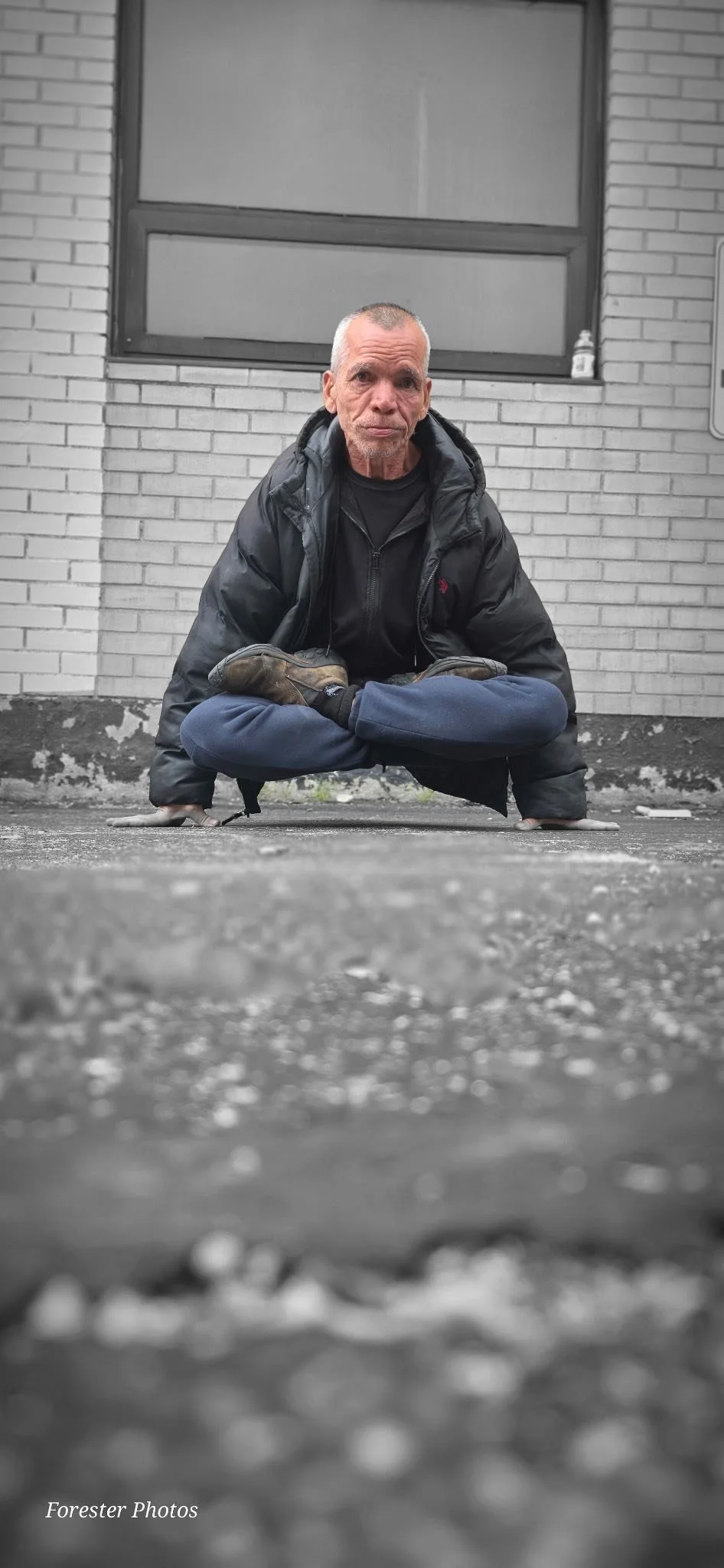 A man with short gray hair and a weathered face, dressed in a black jacket and dark pants, sitting in a lotus position on the ground in front of a gray brick building. The photo is mainly black and white, with the man's clothing and the background in