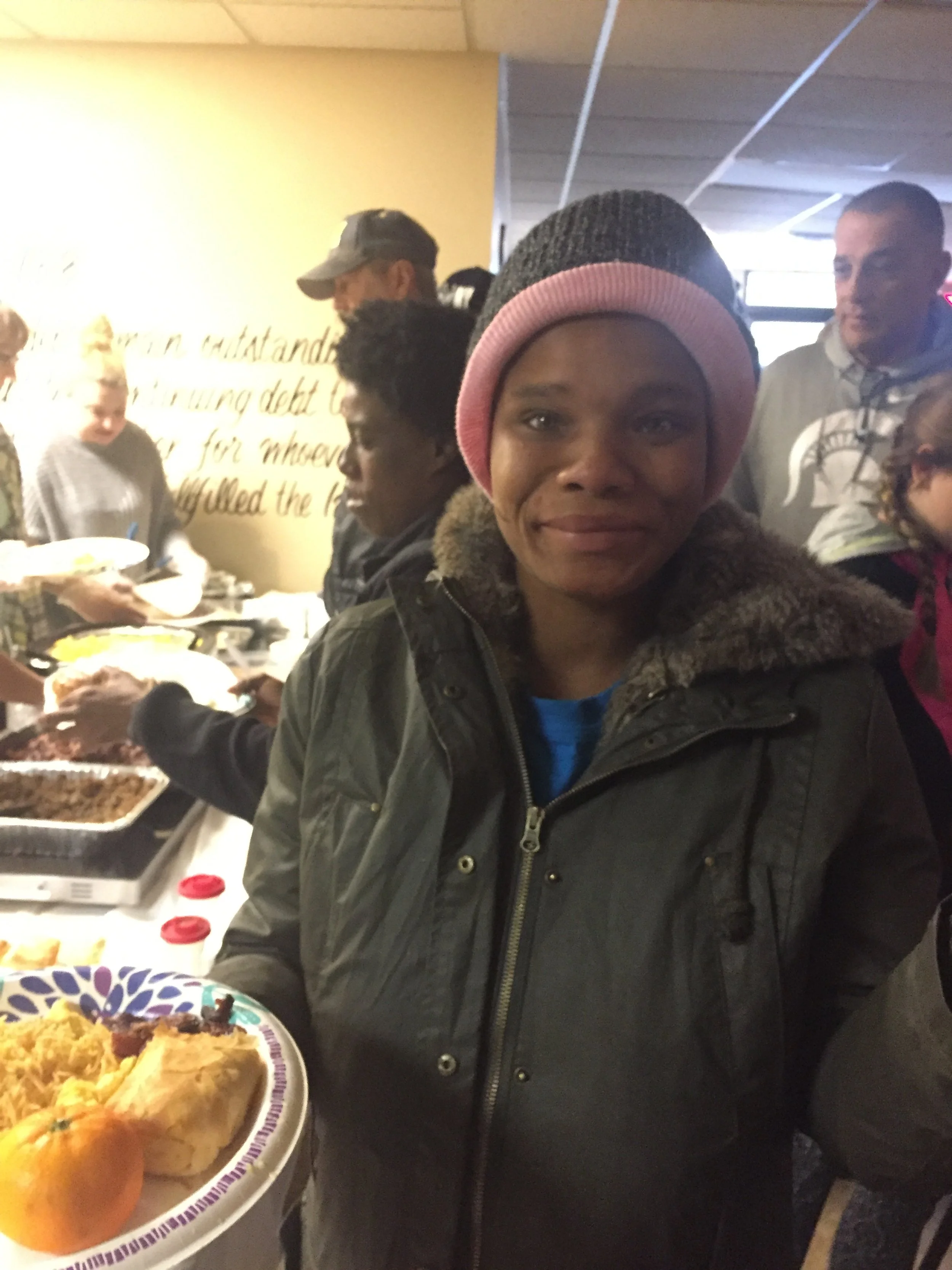 A woman in a warm jacket and pink beanie holding a plate of food at a buffet table with other people in line behind her.