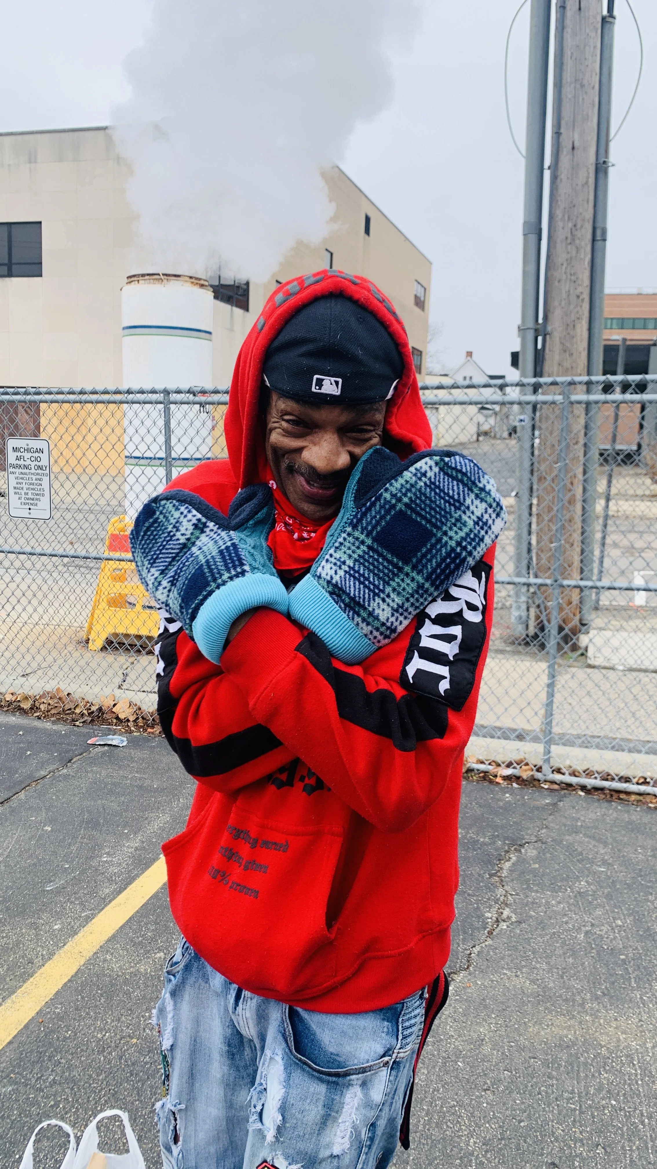 A cheerful man wearing a black baseball cap, red hoodie, and plaid mittens smiling with crossed arms in a parking lot.