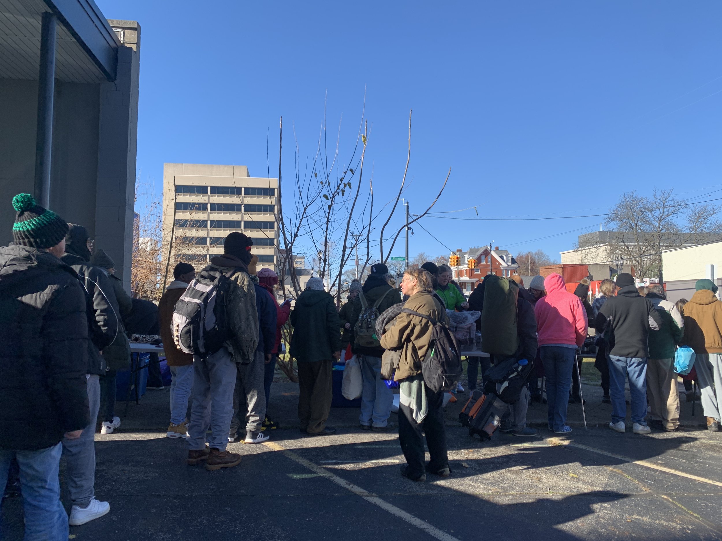 People gathered outdoors in a parking lot on a sunny day, wearing winter clothing, around tables with items, some standing in line or talking, with buildings and leafless trees in the background.