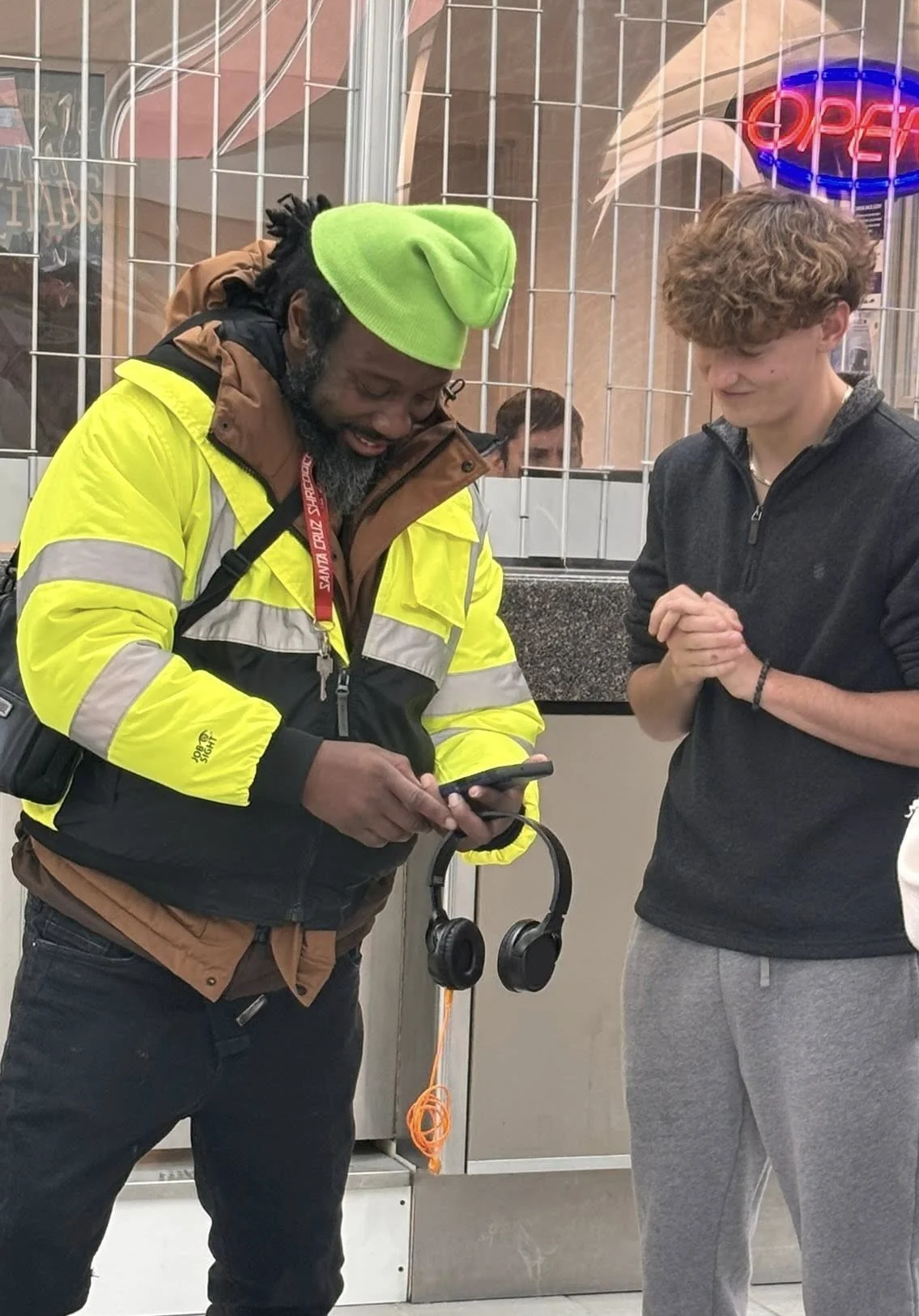 Two men are standing together in an indoor public space, one is showing something on his phone to the other. The man on the left is wearing a yellow jacket, a green beanie, and has headphones hanging from his neck. The man on the right is wearing a black zip-up hoodie and gray sweatpants, with his hands clasped together and smiling.