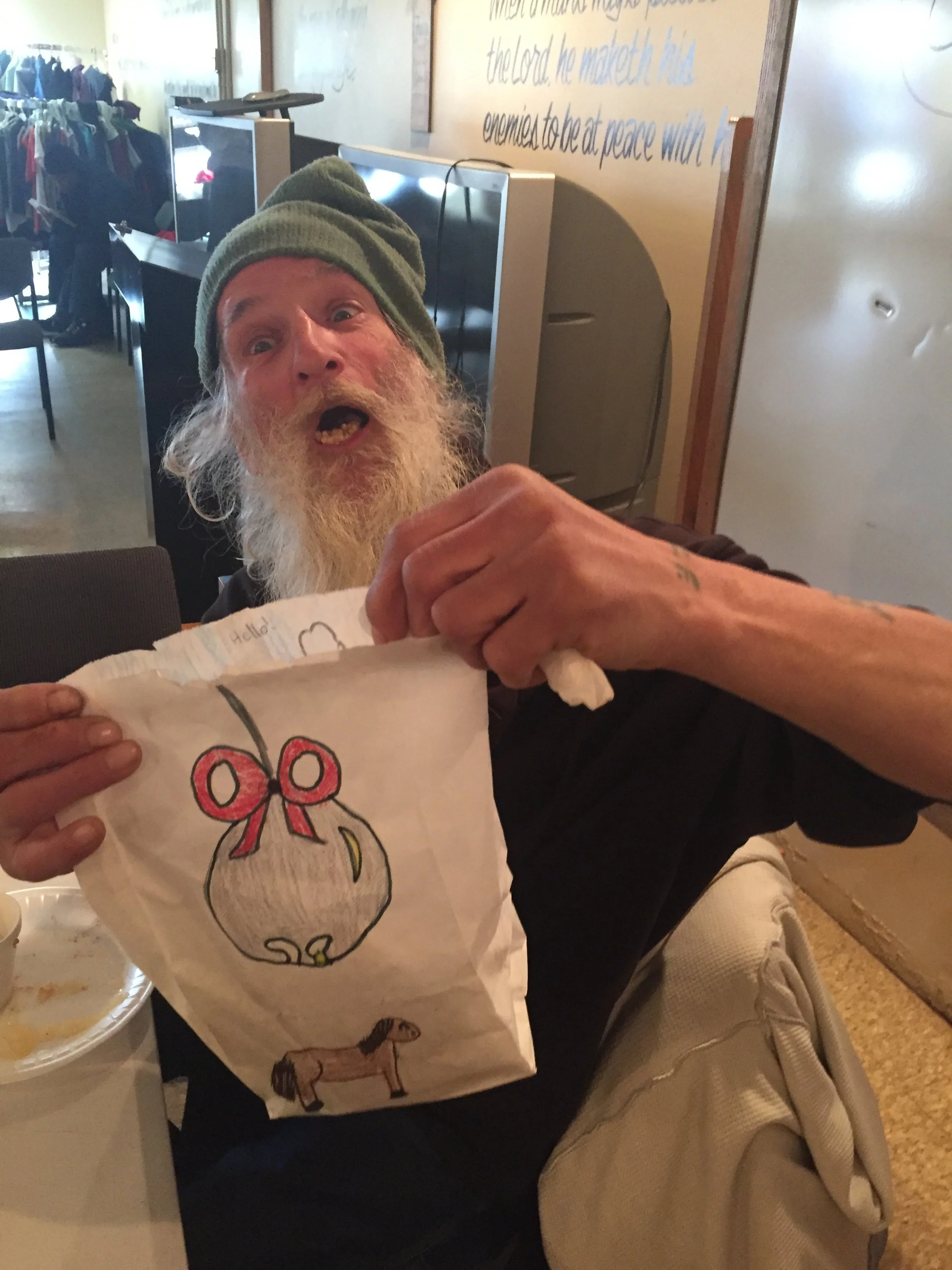 A cheerful old man with a long white beard and green beanie, showing a paper bag with a hand-drawn Christmas ornament and horse, sitting at a table in a restaurant.