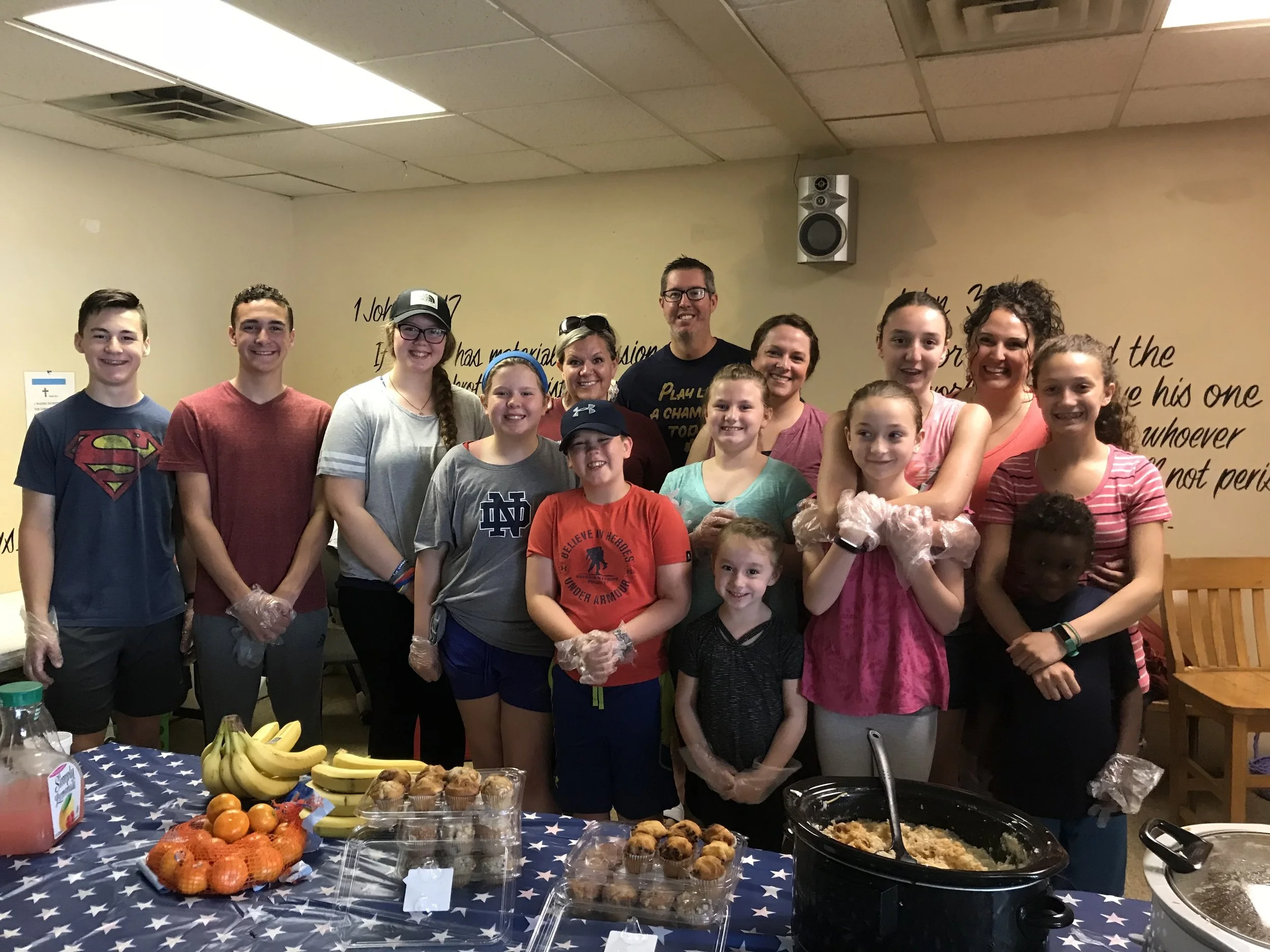 Group of people, including children and adults, smiling behind a table of food in a community or charity setting.
