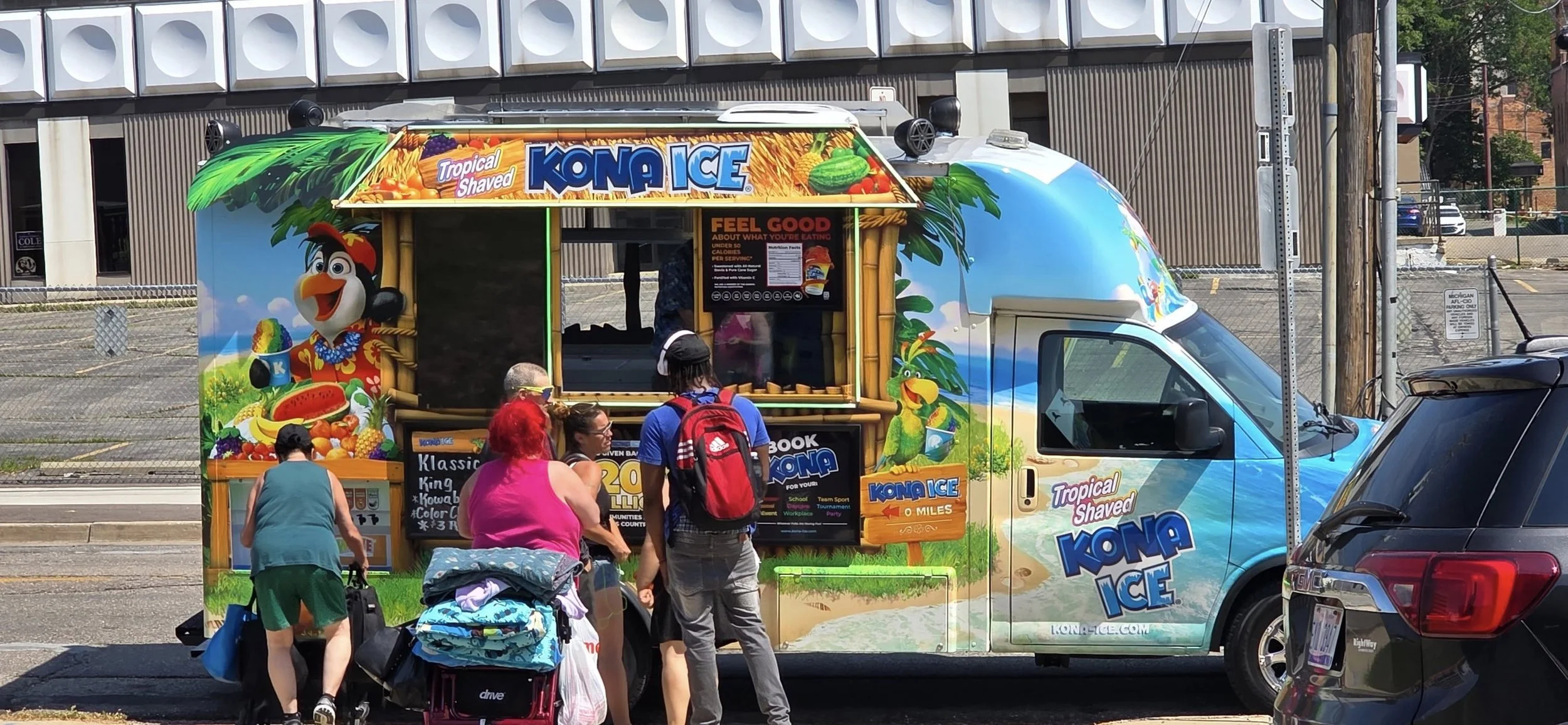 Colorful Kona Ice truck decorated with tropical and beach scene graphics, featuring a penguin mascot, serving customers outdoors.