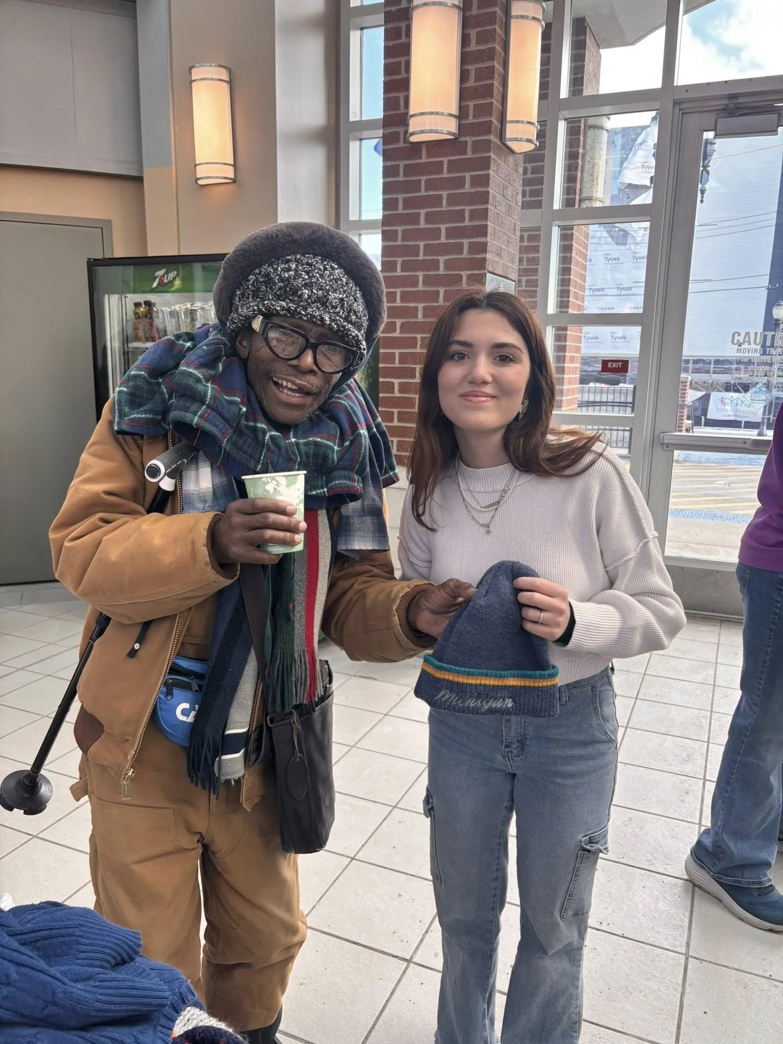 An elderly man and a young woman are standing together inside a store near a glass door. The man is smiling, wearing glasses, a winter hat, a scarf, a brown jacket, and holding a cup. The woman is smiling, wearing a cream sweater and holding a blue winter hat, standing with her arm around the man.