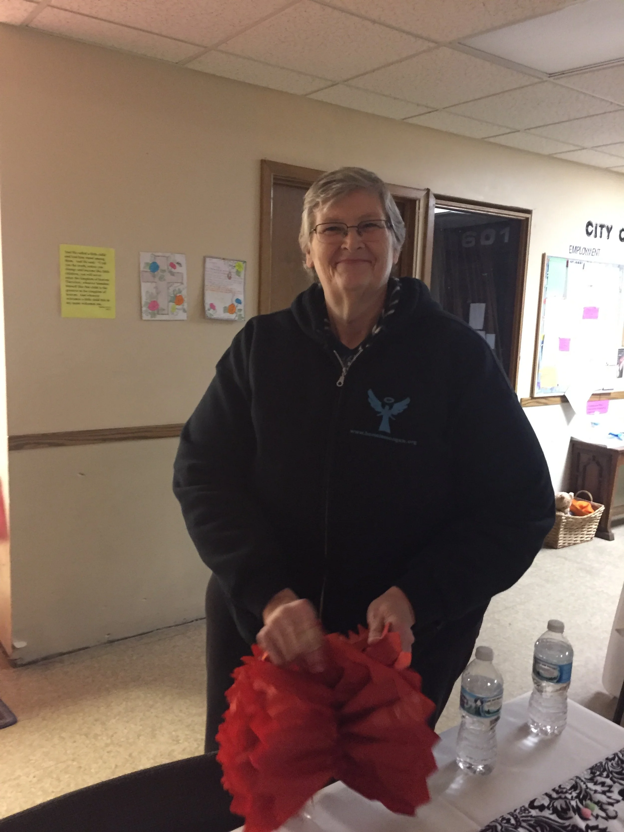 A smiling older woman with short gray hair and glasses, wearing a black hoodie with a blue logo, standing indoors near a table with water bottles and red decorative paper.