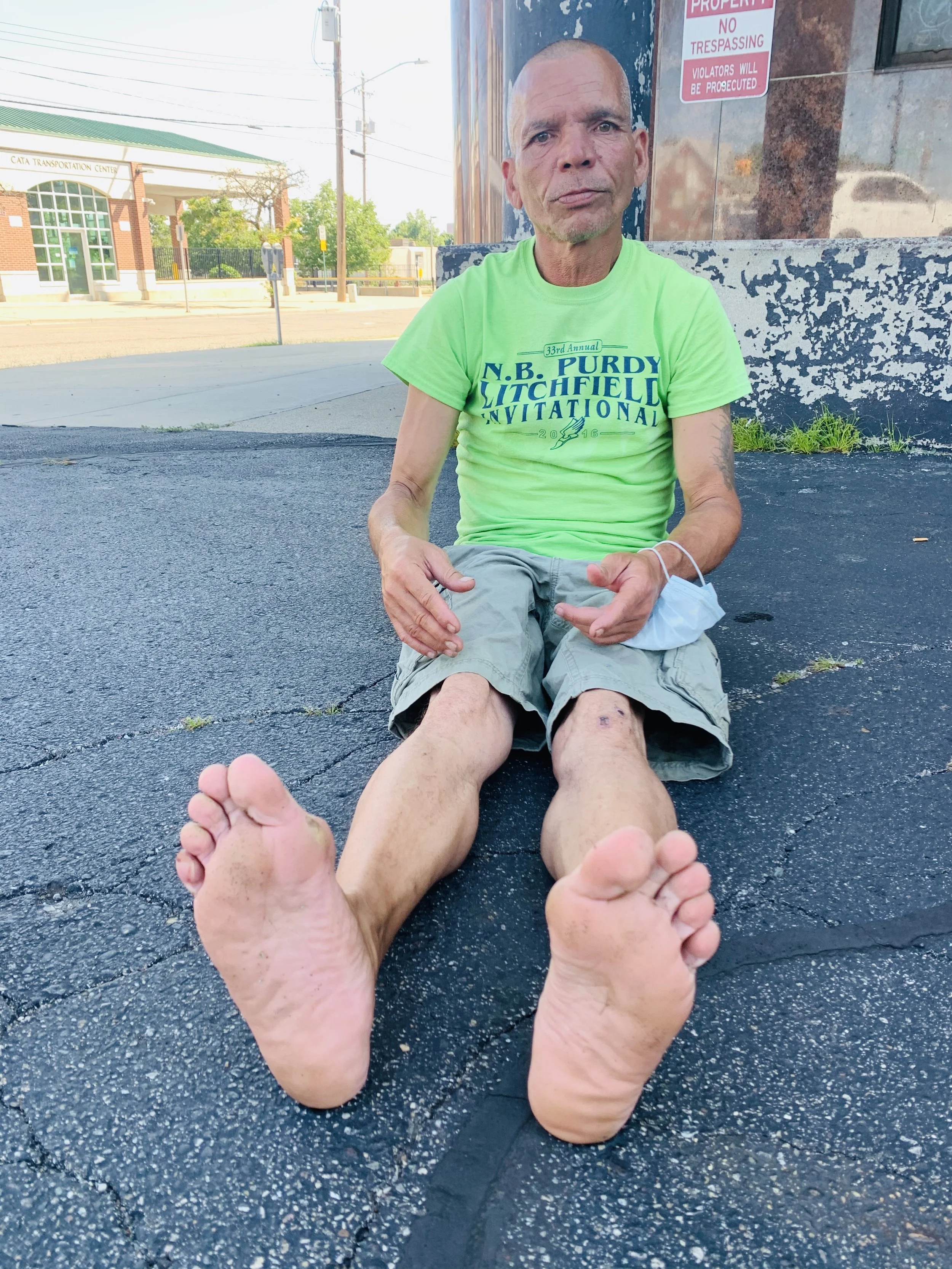 A man sitting on the sidewalk with dirty feet, wearing a bright green T-shirt and khaki shorts, holding a face mask, with a brick building and street in the background.