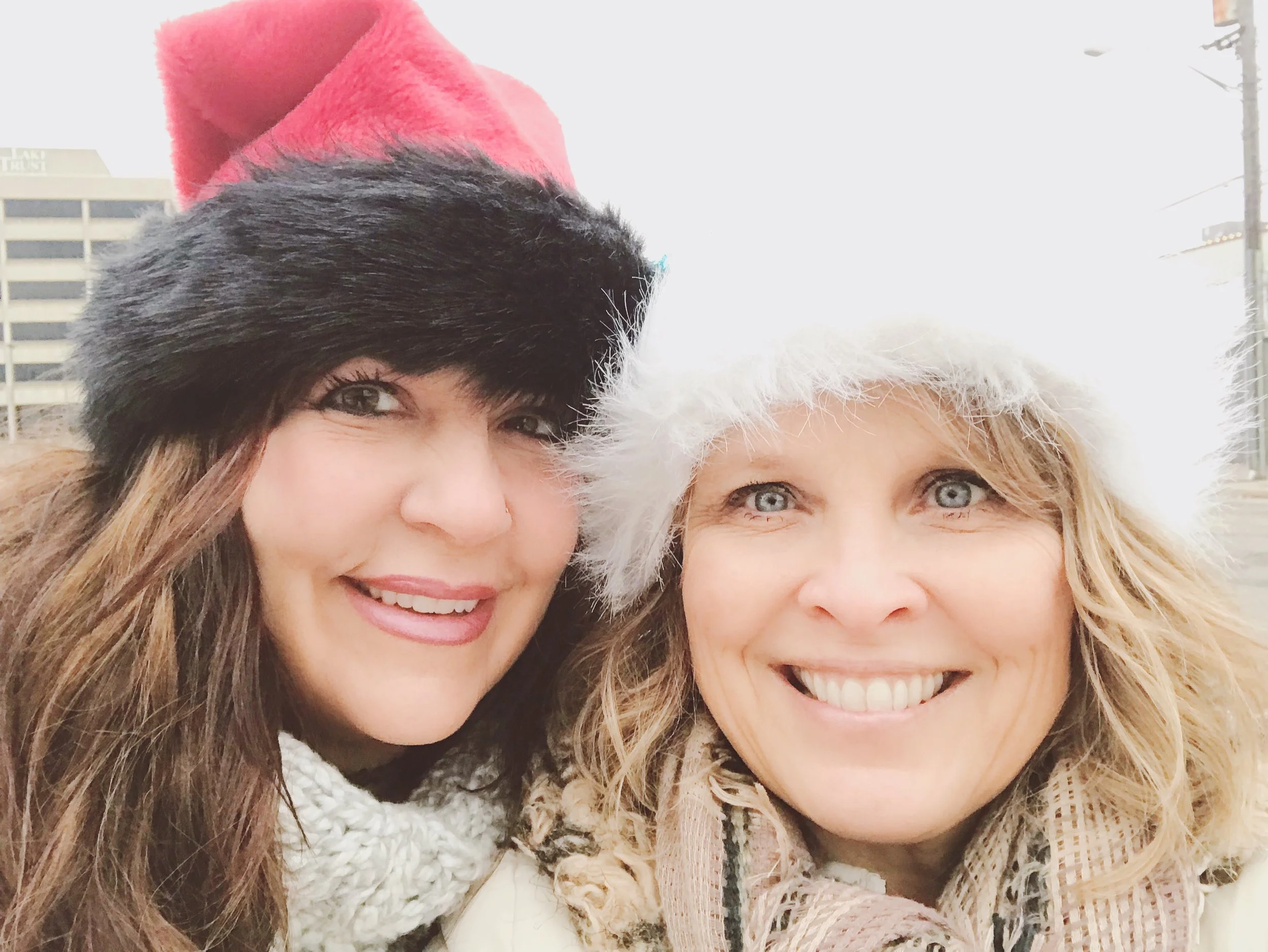 Two women smiling outdoors in winter, wearing festive hats and scarves.