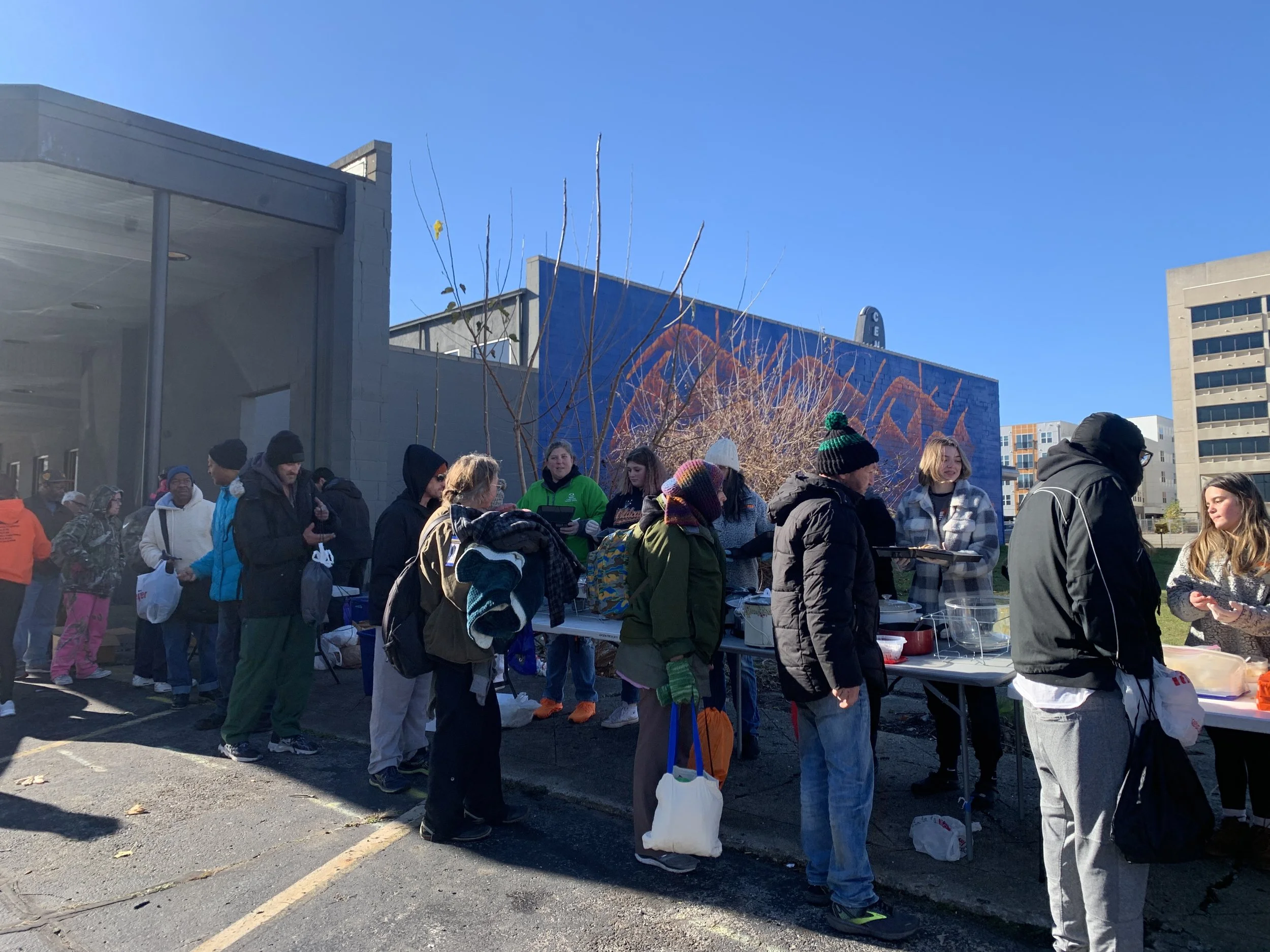 People line up outside a building on a sunny day, receiving food or goods from tables set up along the sidewalk, with an urban backdrop of colorful buildings and a mural.