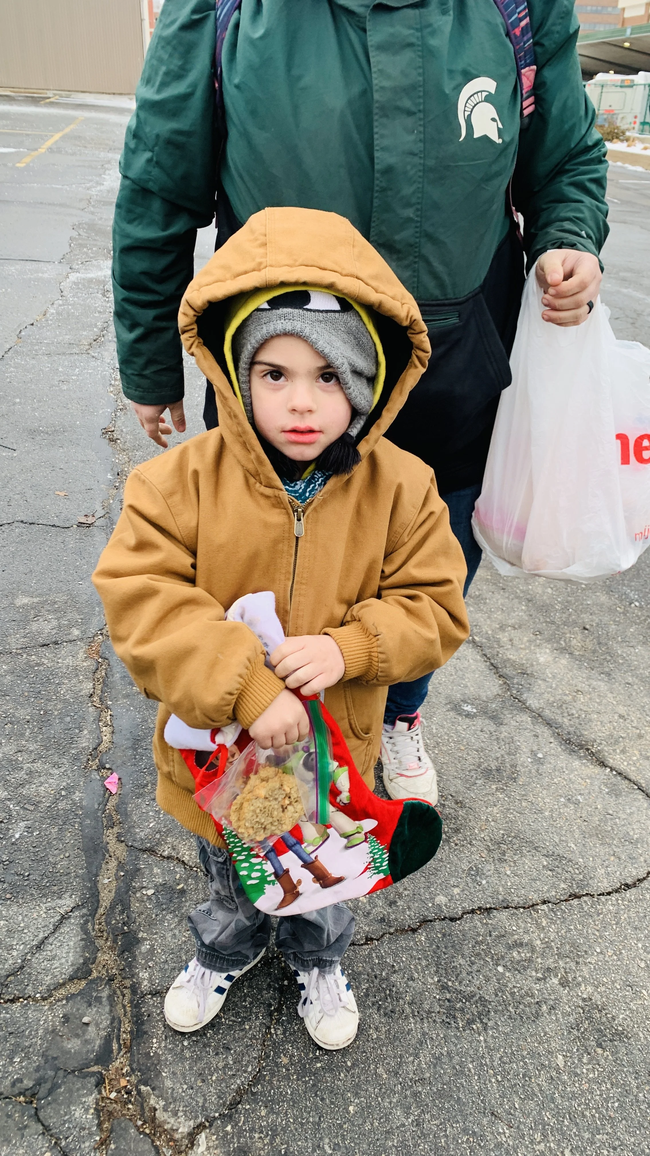 A young boy wearing a brown hooded jacket, gray and yellow beanie, and gray pants stands on a cracked pavement holding a Christmas-themed gift bag with a cookie inside. Behind him, an adult wearing a green jacket with a Michigan State Spartan logo an