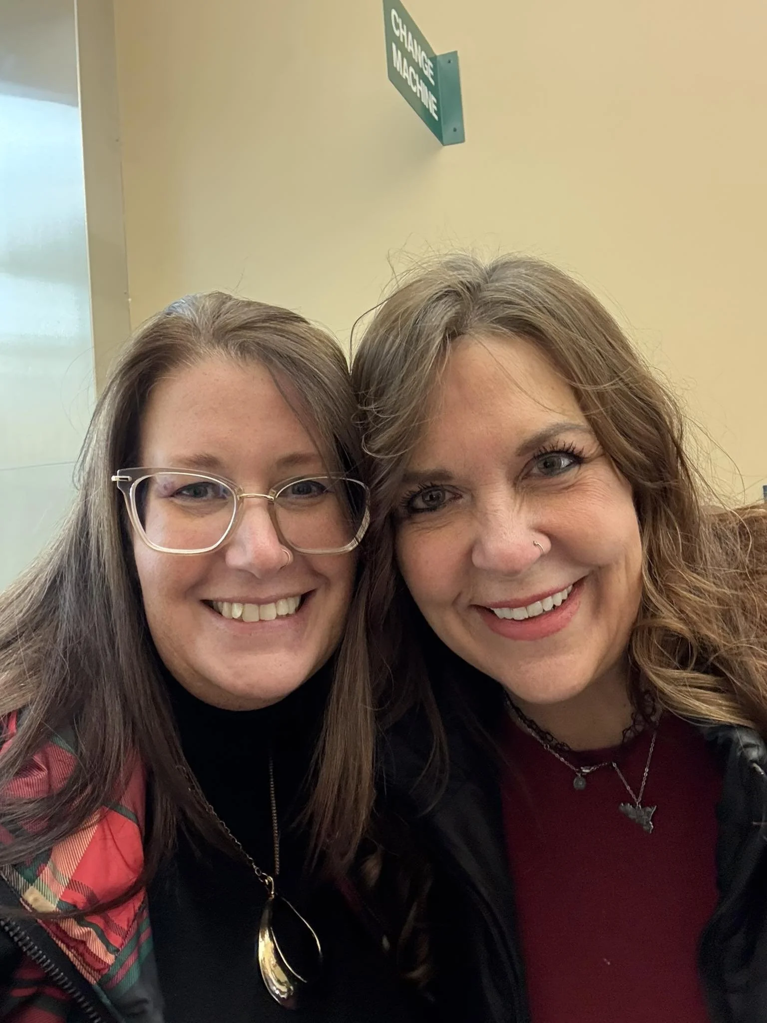 Two women smiling for a selfie indoors, with a sign in the background that reads 'Change Machine'.