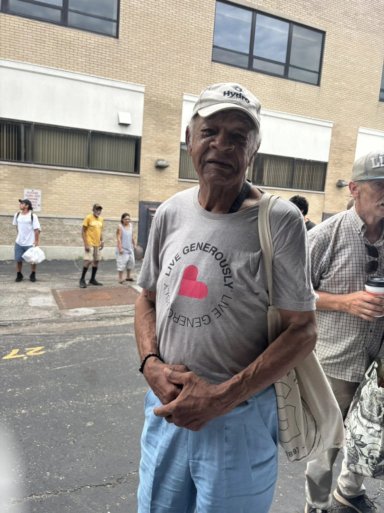 An elderly man standing outdoors on a city street, wearing a gray t-shirt with a pink heart and text, light blue shorts, a beige cap, and a black bracelet. Other people are visible in the background.