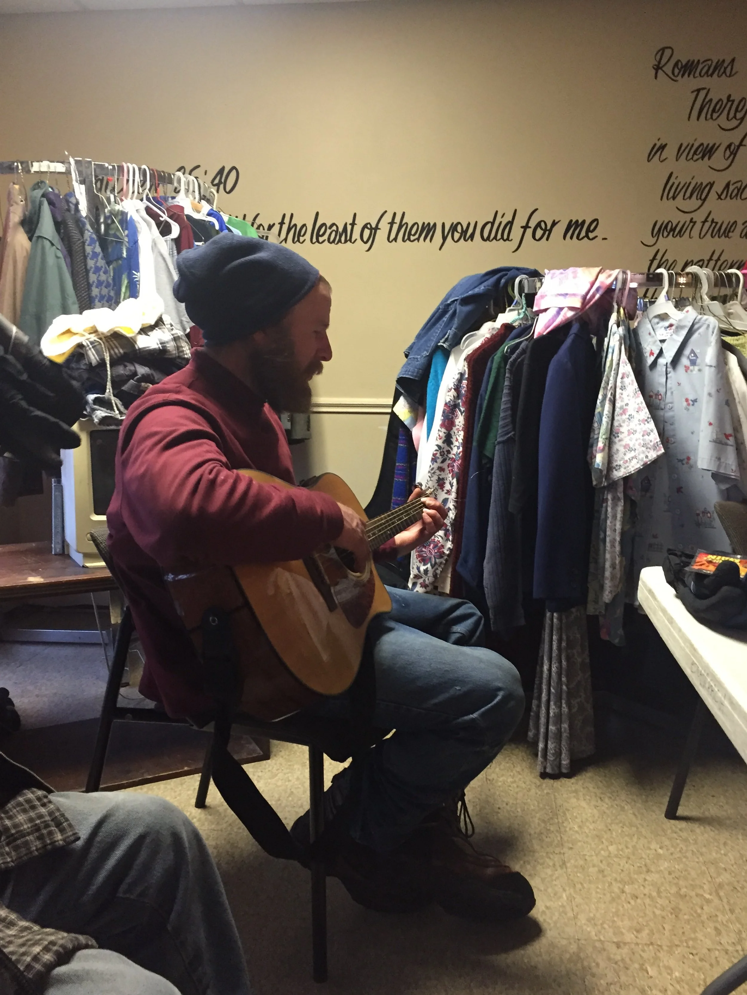 A man in a maroon jacket and a navy beanie playing an acoustic guitar in a cluttered room filled with hanging clothes and a small desk and chair.