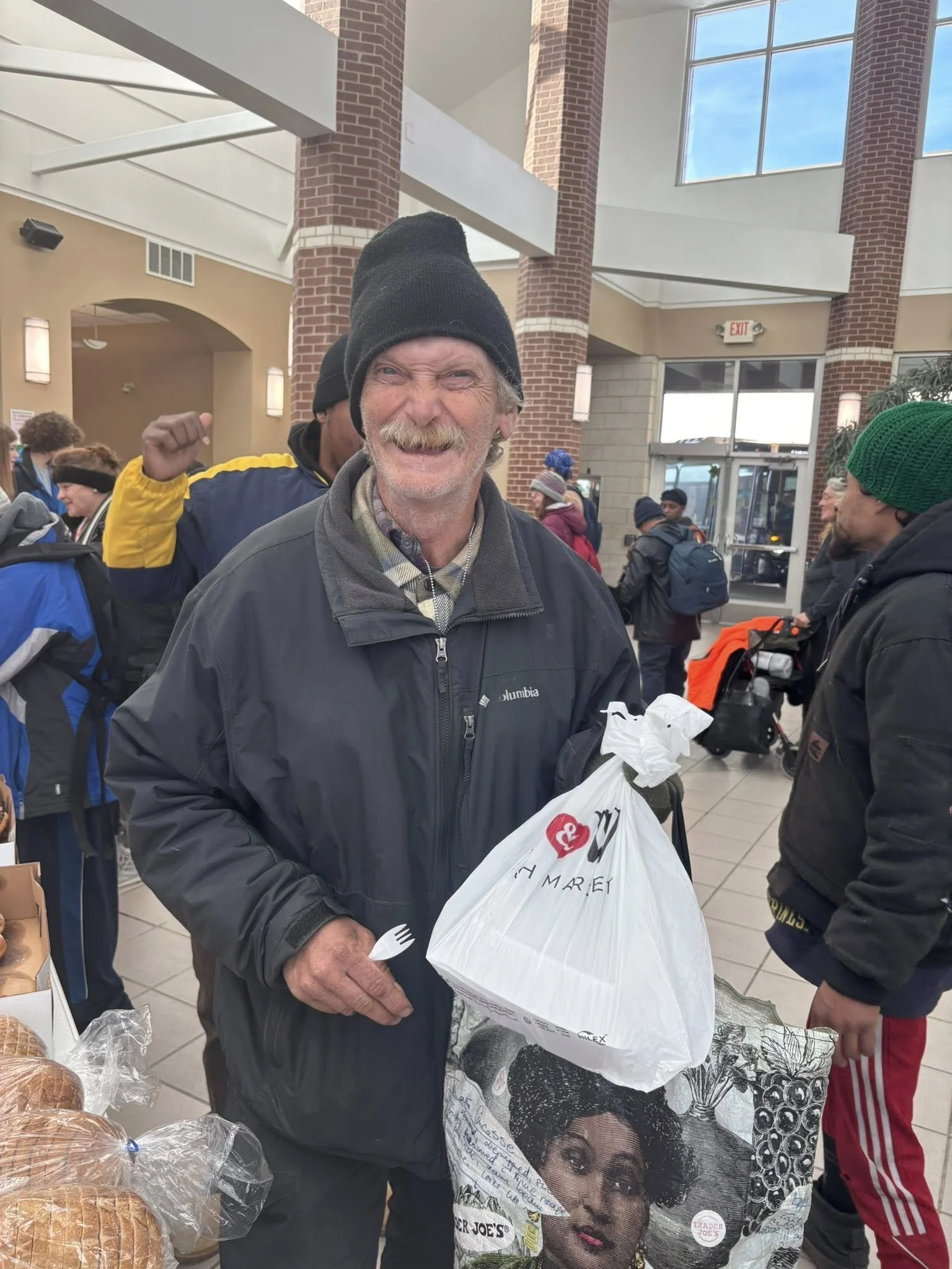 A smiling older man wearing a black beanie and black jacket holding a shopping bag inside a busy indoor marketplace or grocery store.