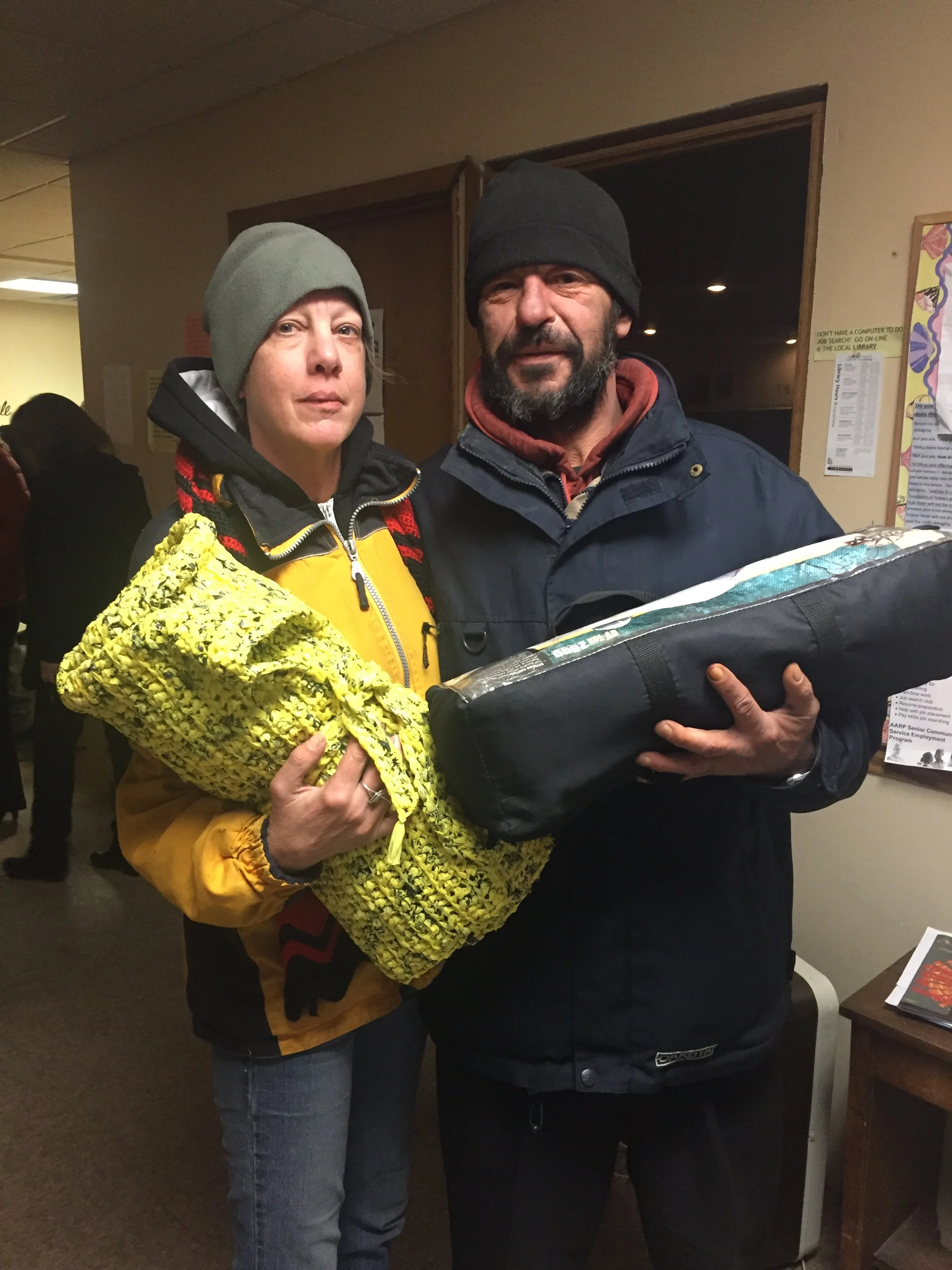 A woman and a man standing indoors, holding a rolled-up item in a yellow, textured bag and a black bag. Both are wearing dark jackets and beanies, and the woman is wearing a yellow jacket underneath. There are other people and posters visible in the 
