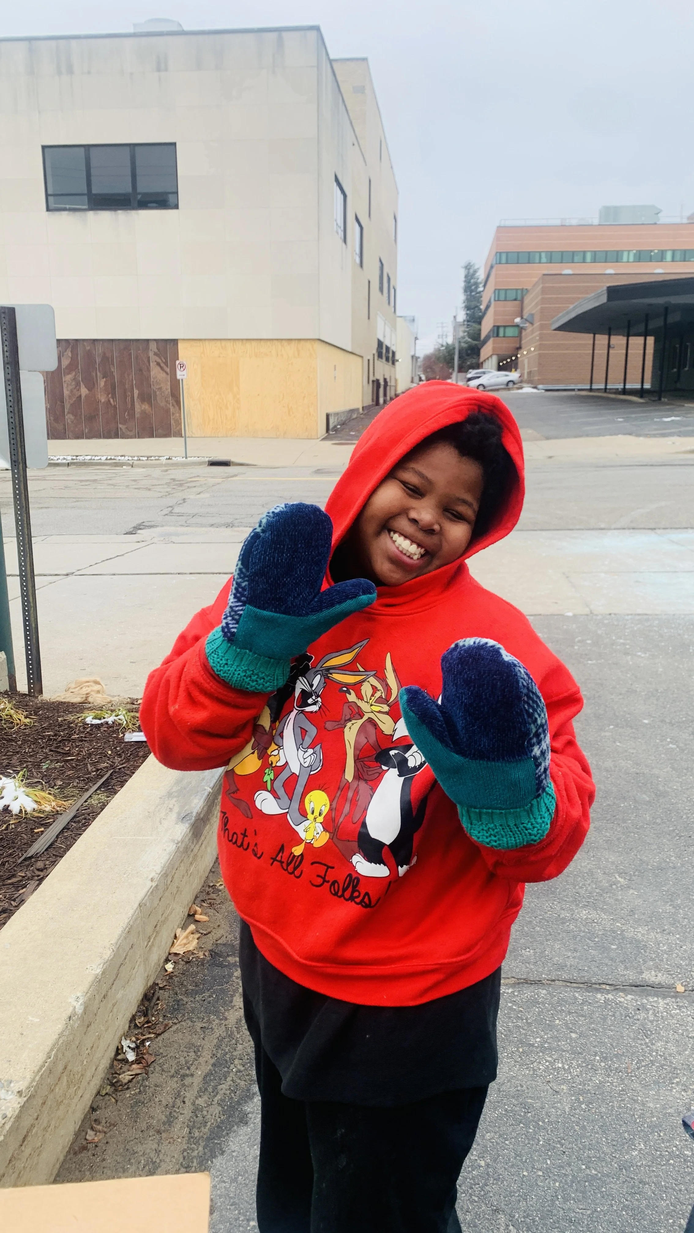 A smiling child wearing a red hoodie with Looney Tunes characters and gloves, standing outdoors on a sidewalk.