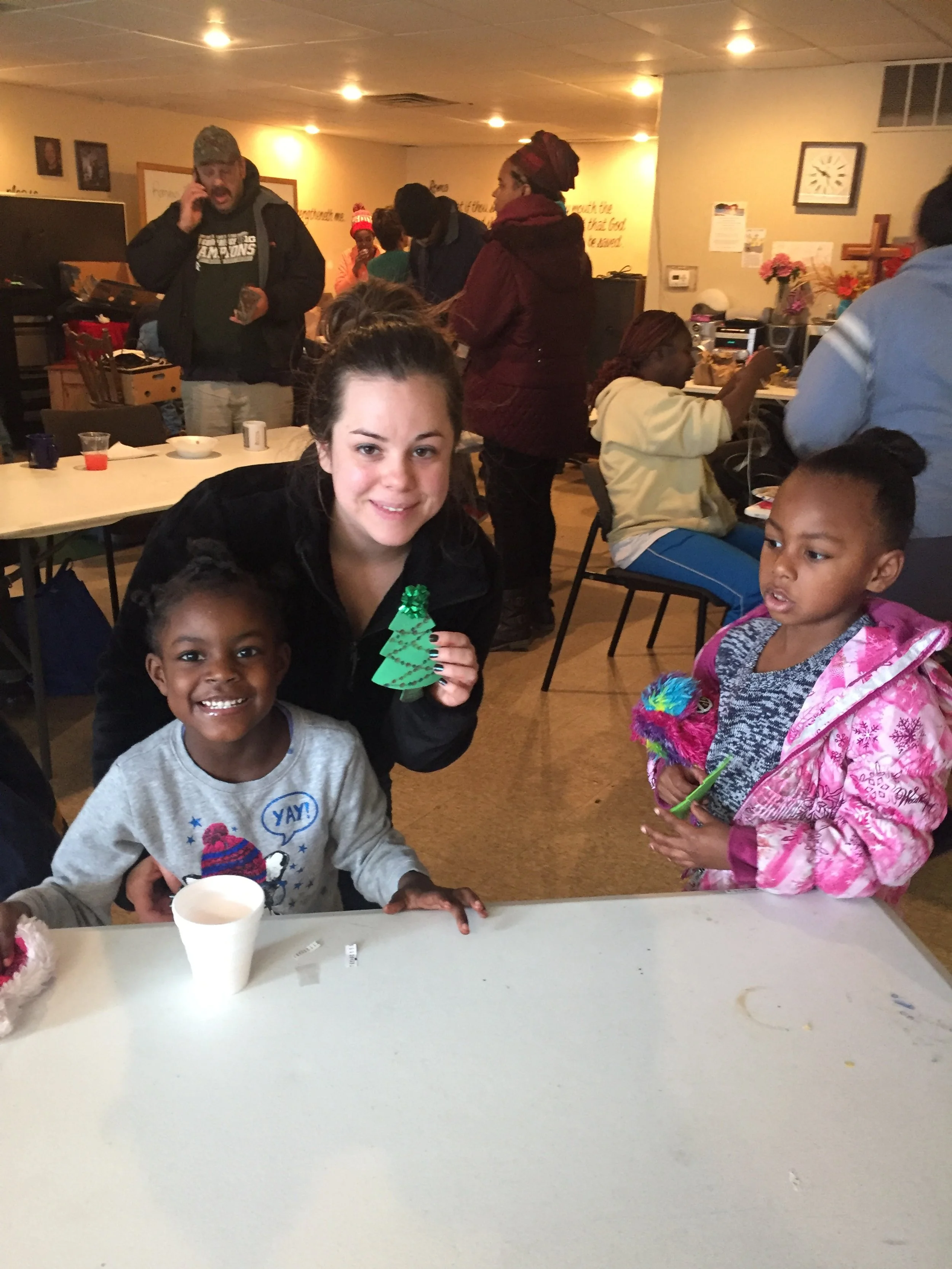 A group of children and adults gathered in a room for a celebration, with two young girls smiling at the camera, one holding a small green Christmas tree decoration, and the other holding a colorful toy. The background shows people socializing, table