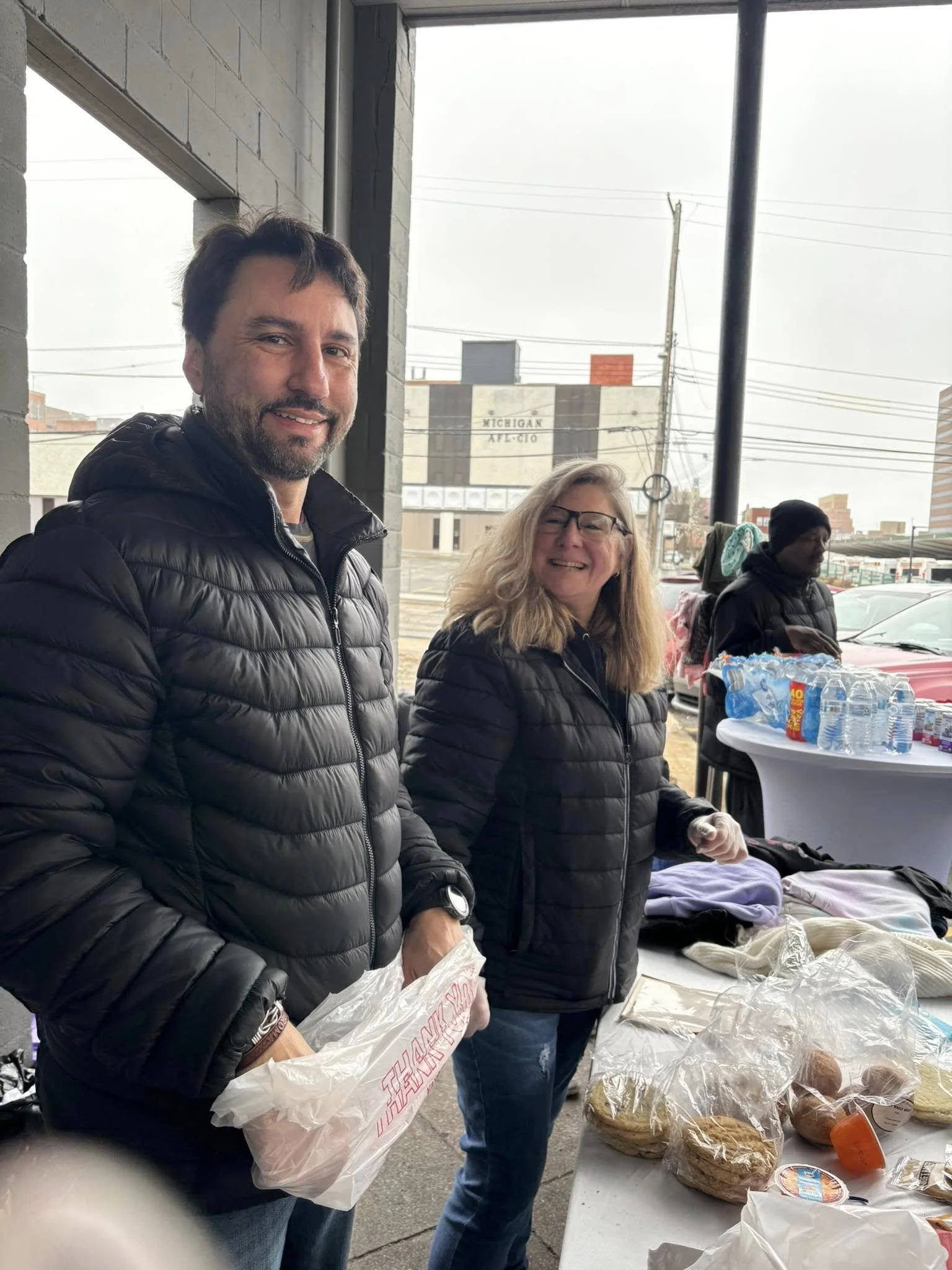 Two people smiling at a table with baked goods and supplies, outdoors on a cloudy day.