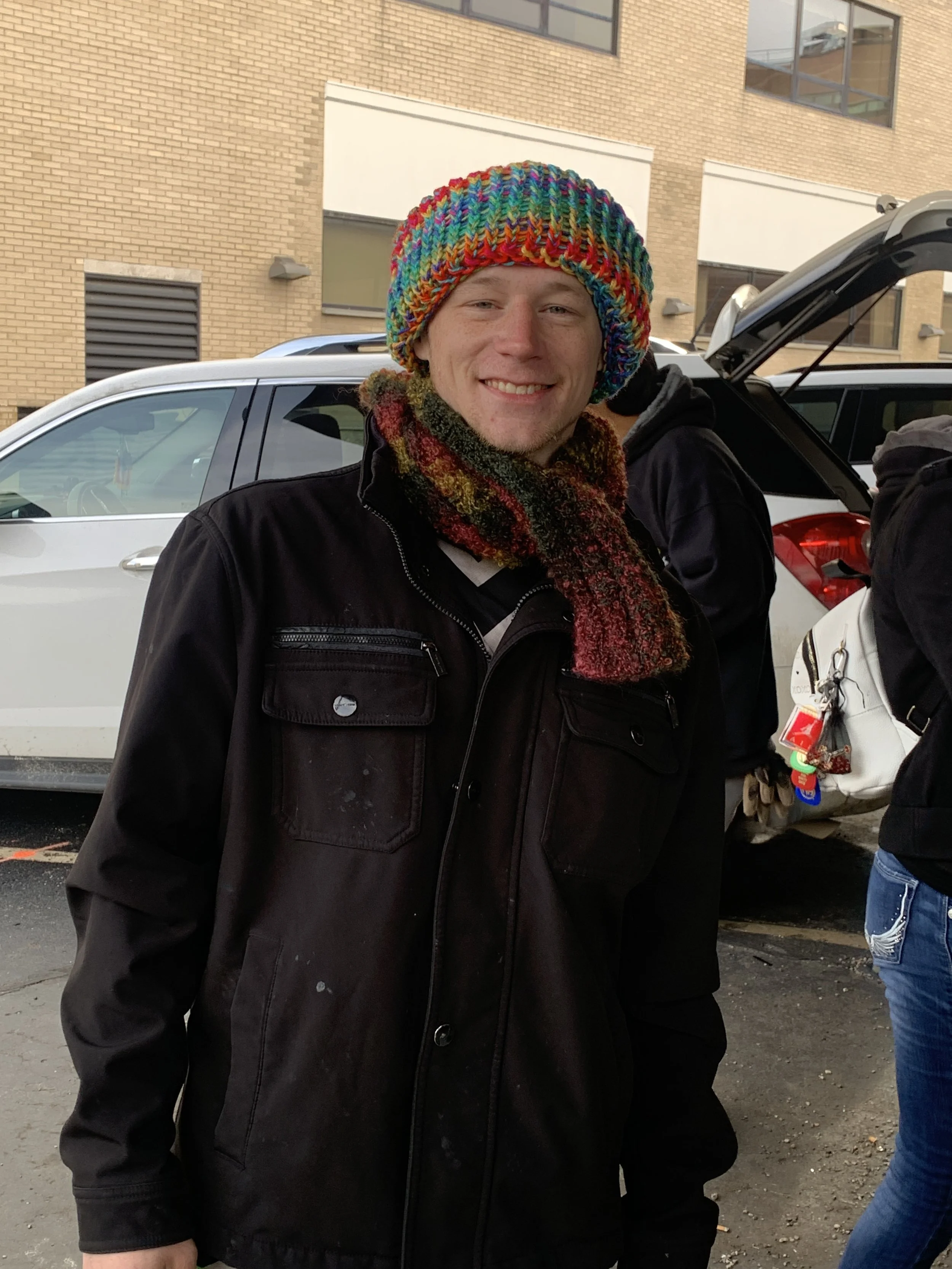 A young man smiling wears a multicolored knit hat and a scarf, standing outdoors in front of parked cars and a brick building.