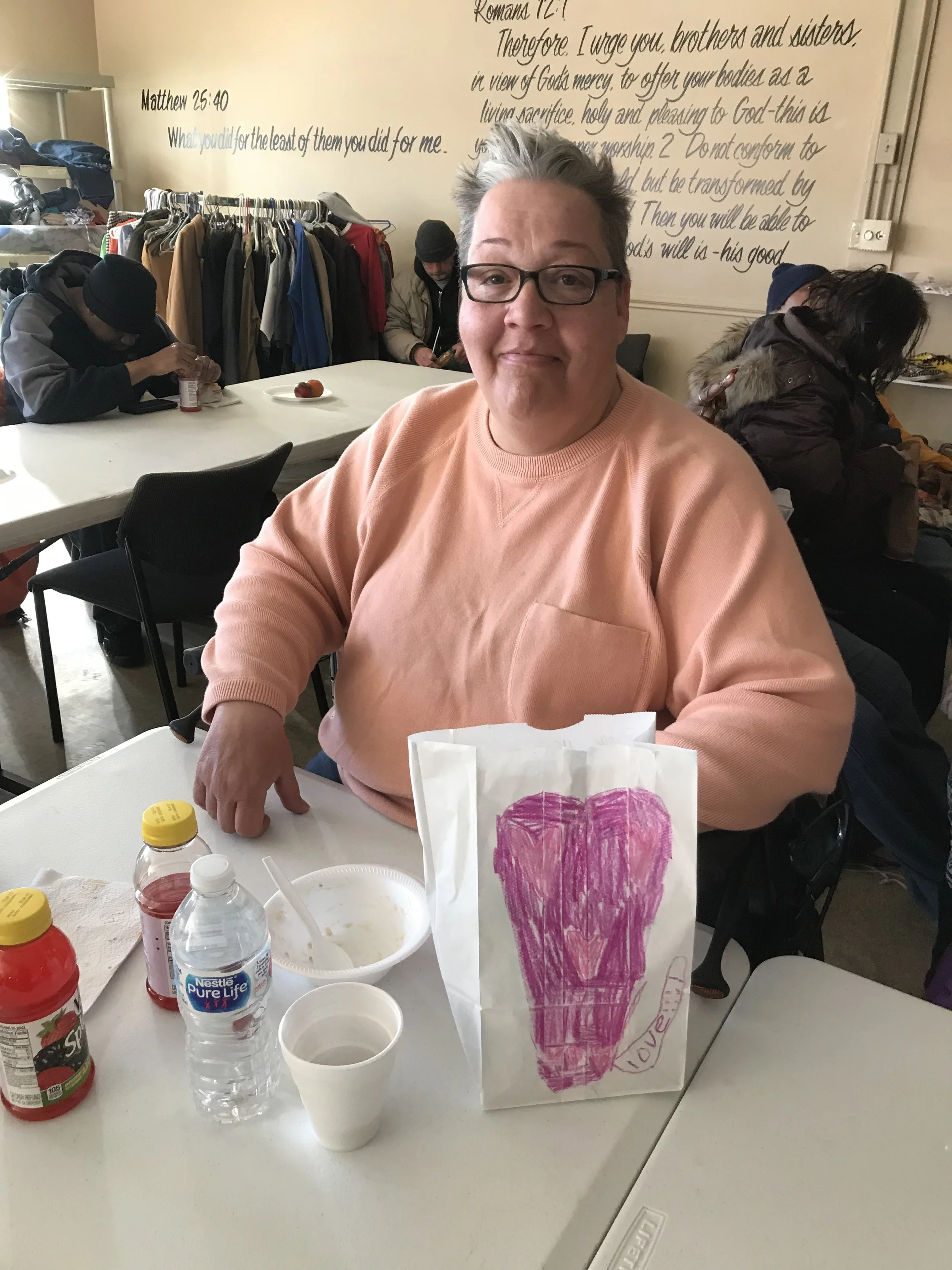 A woman with short gray hair, glasses, and a peach-colored sweatshirt sitting at a table with a paper bag decorated with a pink heart and the word 'LOVE,' along with a water bottle, juice bottles, a small cup, and a bowl. In the background, people ar