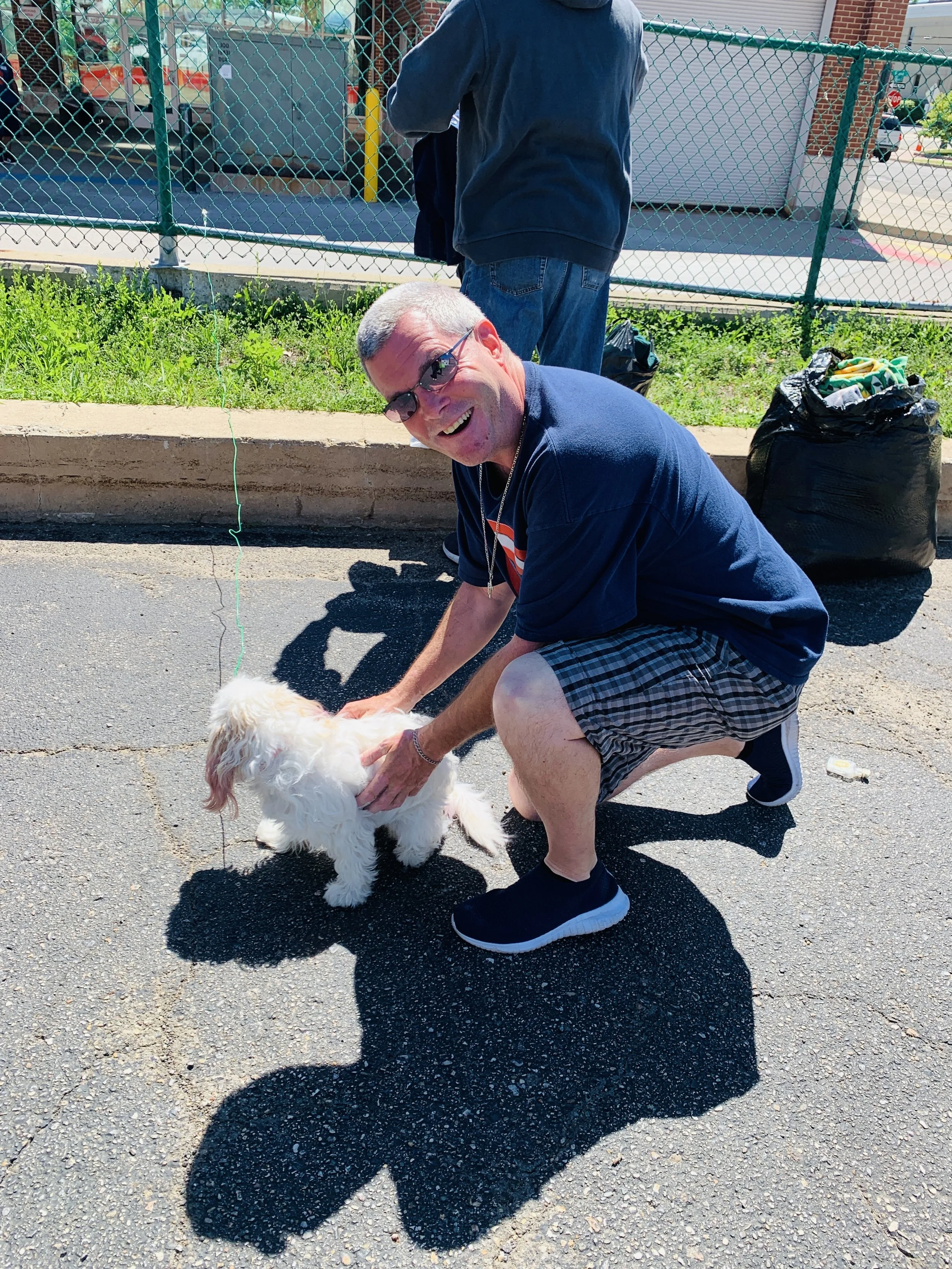 A man crouching on the street petting a small white dog with long ears. The man is wearing glasses, a black t-shirt, plaid shorts, and slip-on shoes. Behind them, a person with their back turned stands near a fence. There are green grass and bags of 