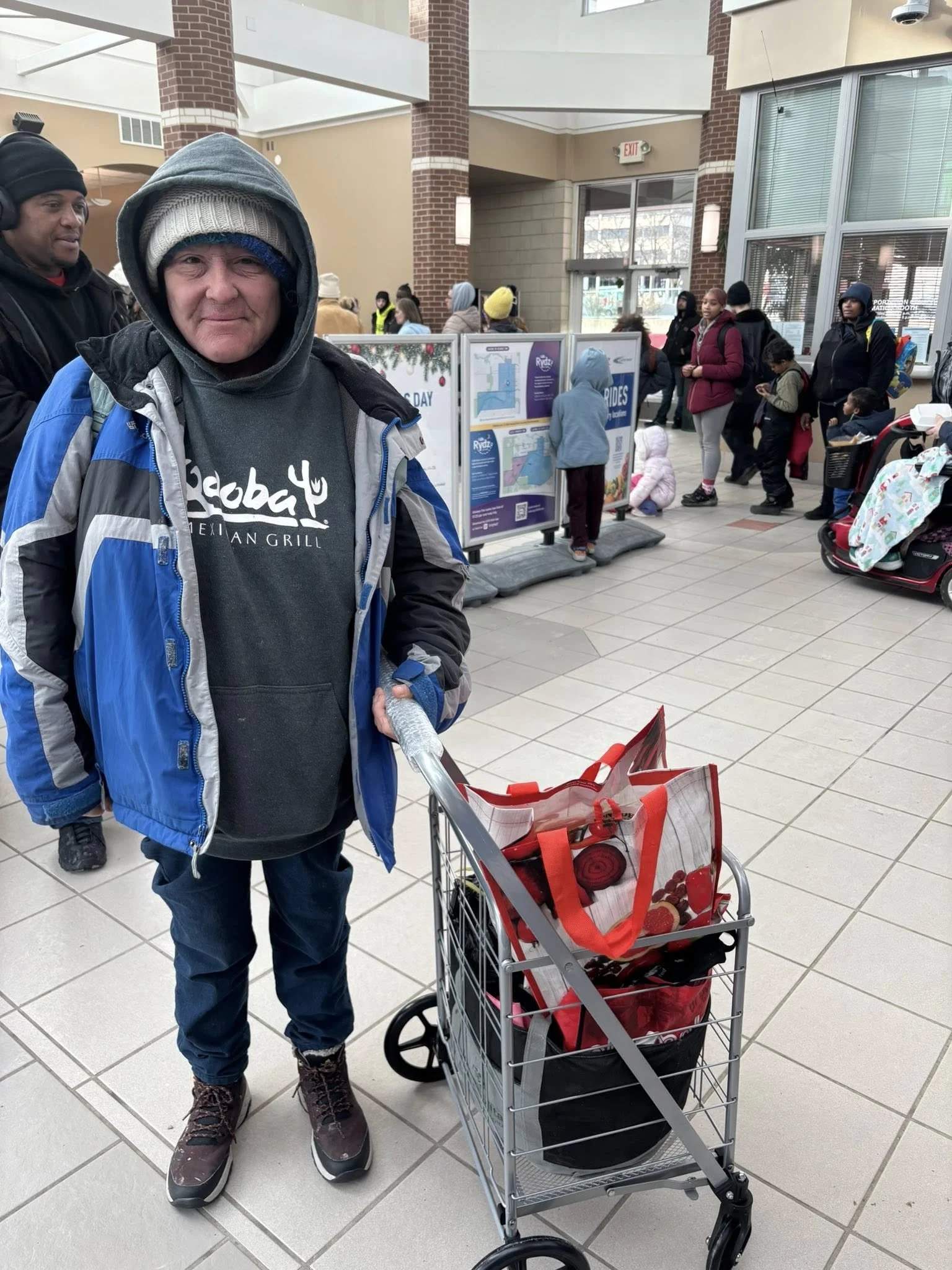 An older man in winter clothing standing next to a shopping cart inside a busy store or mall. He is wearing a gray hoodie under a blue and gray jacket, and has a gray beanie with a blue hat underneath. The shopping cart contains a red tote bag and ot