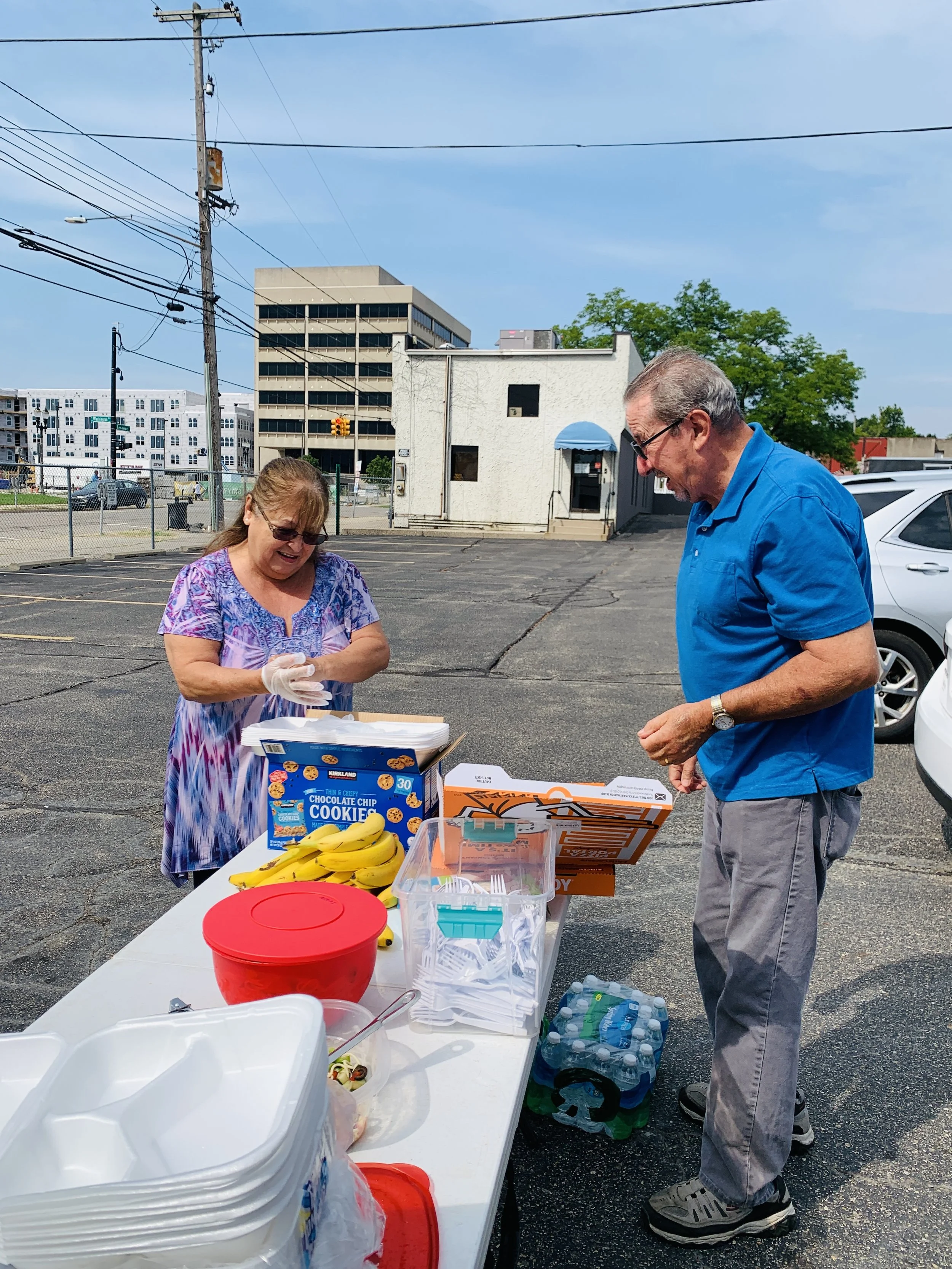 A woman and a man are standing at a table with food, snacks, and bottled water in a parking lot on a sunny day. The woman is wearing glasses and a colorful dress, while the man is dressed in a blue polo shirt and gray pants.