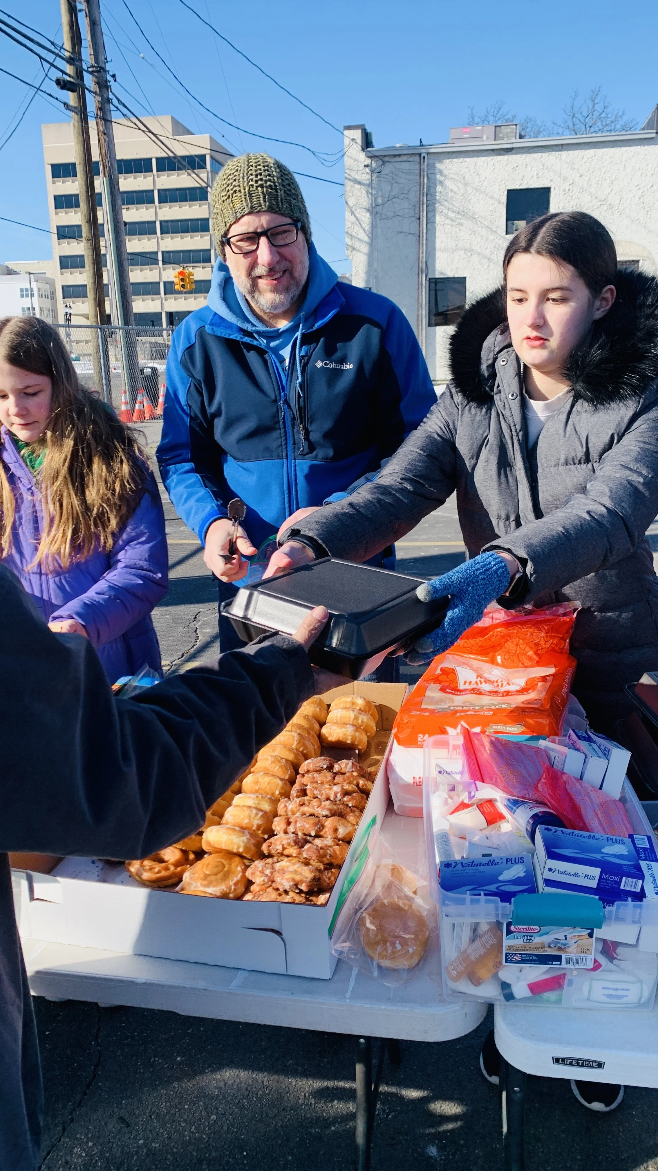 A man with glasses and a beanie wearing a blue jacket is handing a food container to a woman in a gray coat. Two young girls are standing nearby, one in a purple coat and the other not clearly visible. They are at an outdoor table with donuts, snacks