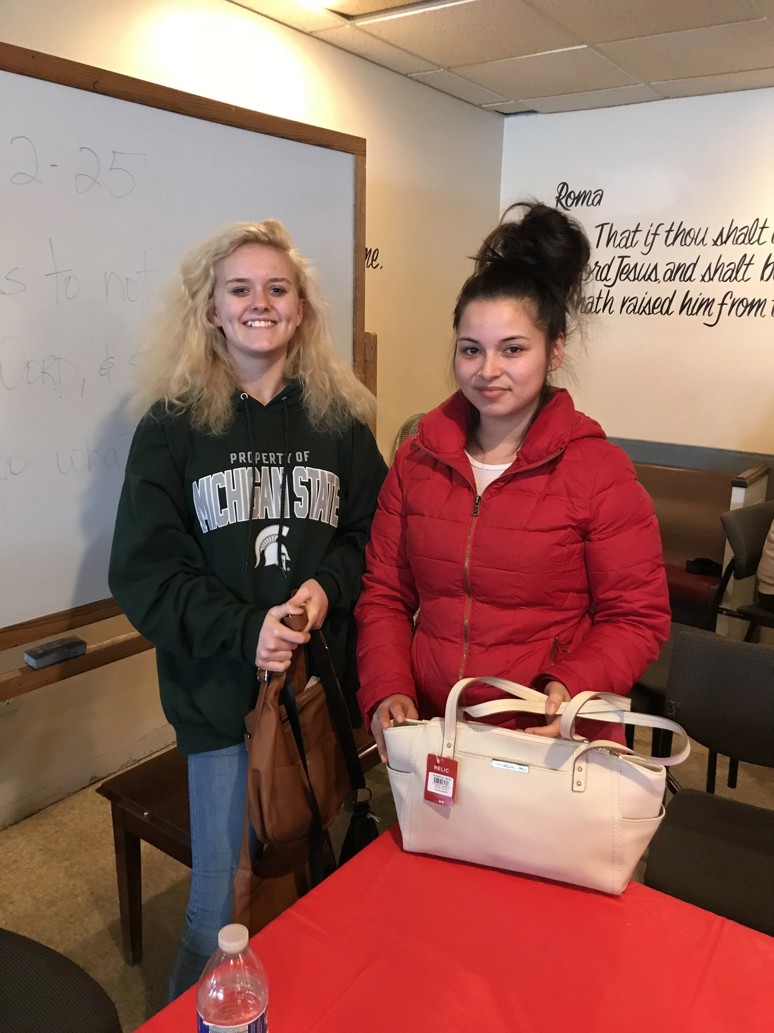 Two young women standing indoors in front of a whiteboard and wall with religious text. One woman has curly blonde hair, wears a black Michigan State hoodie, and is smiling. The other woman has dark hair tied up in a high bun, wears a red jacket, and