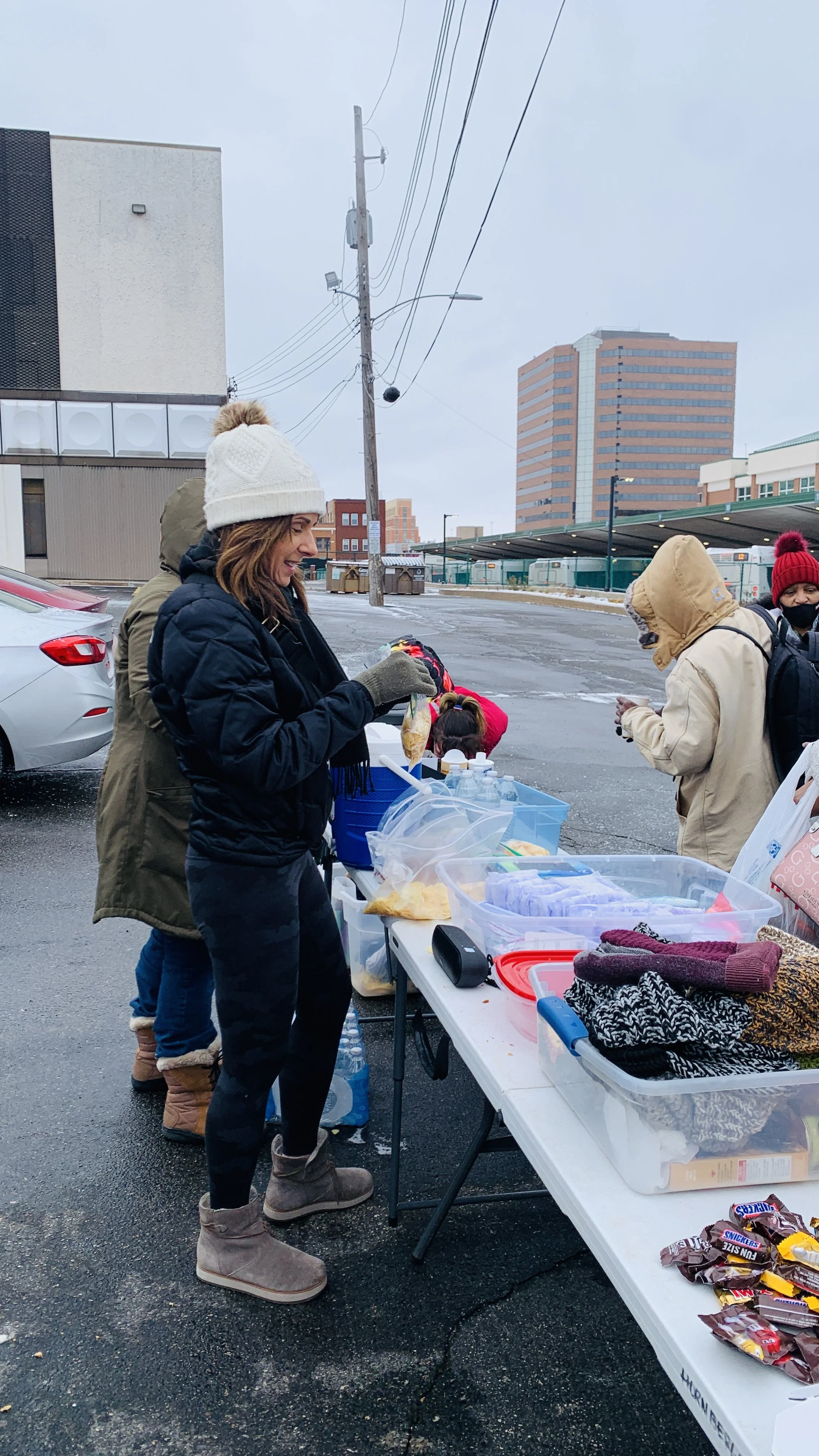 People in winter clothing at outdoor table with food and supplies on a chilly, overcast day in an urban area.