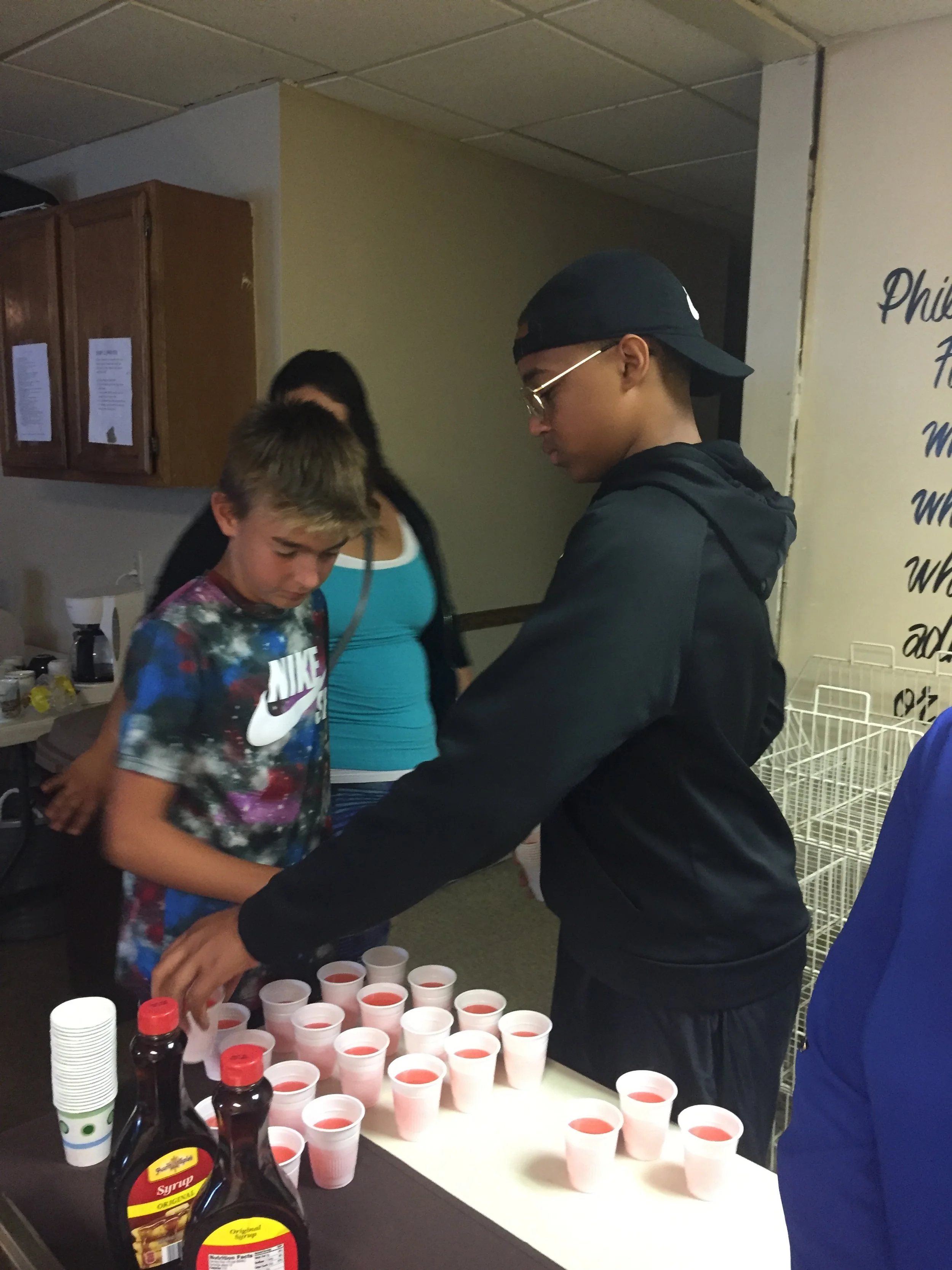 A young man wearing glasses, a black hoodie, and a backwards cap is serving pink-colored drinks in white cups on a table. There is a boy with blonde hair and a colorful Nike shirt standing to the left of him, and a woman in blue in the background. Th