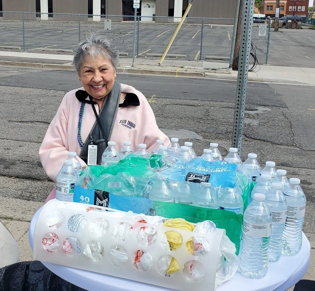Smiling elderly woman standing behind a table with many bottled waters and a plastic carrier for bottles on a sidewalk in an urban area.