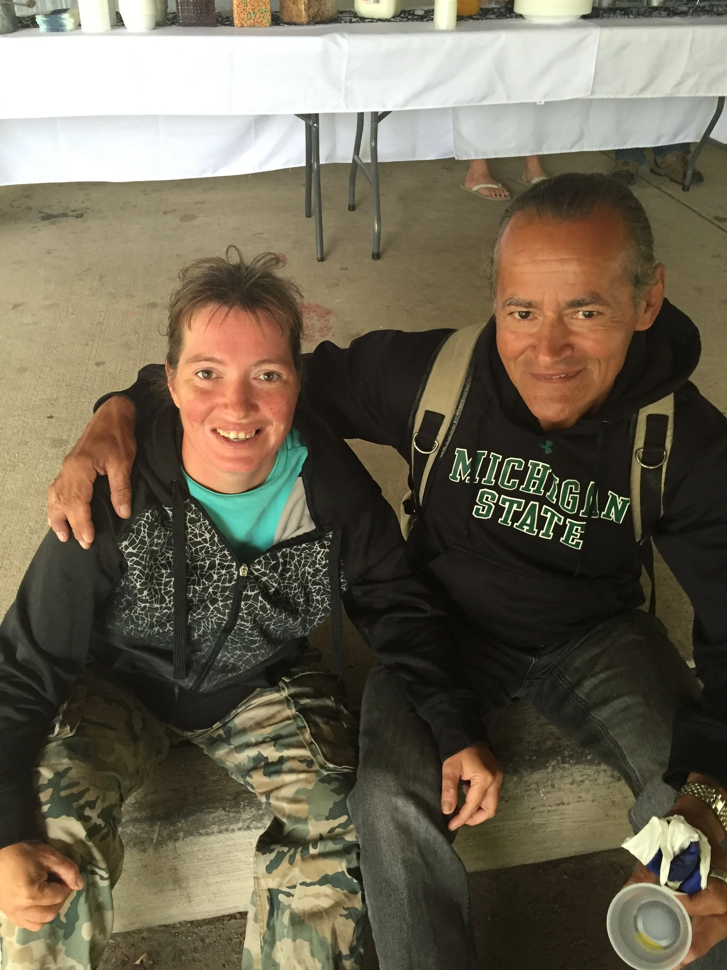 Two men sitting on the ground, smiling at the camera, with one wearing a black hoodie with 'Michigan State' written on it. There are tables with white tablecloths and bottles in the background.