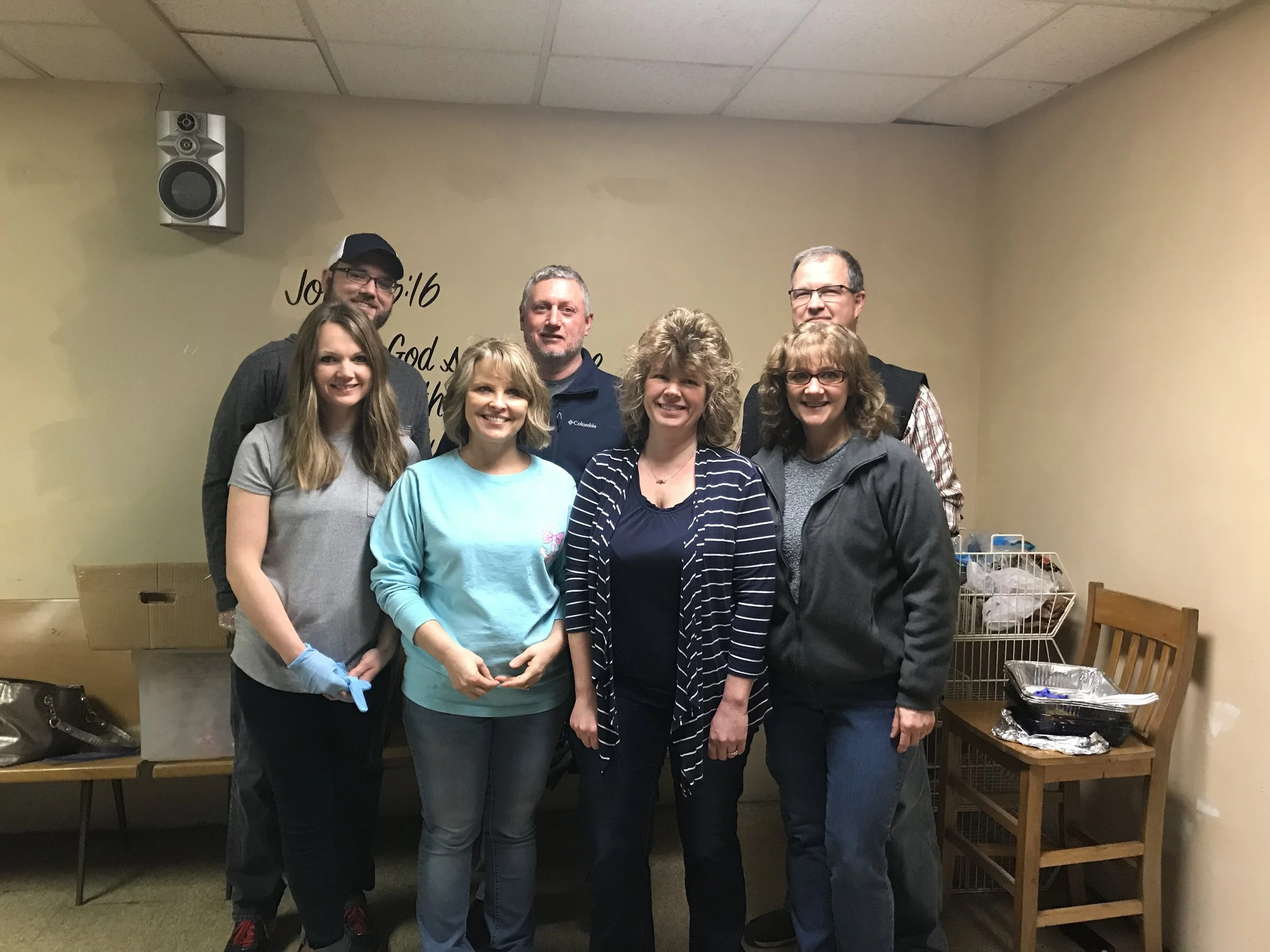 Seven people standing together indoors, smiling at the camera, with a wall behind them displaying a Bible verse.
