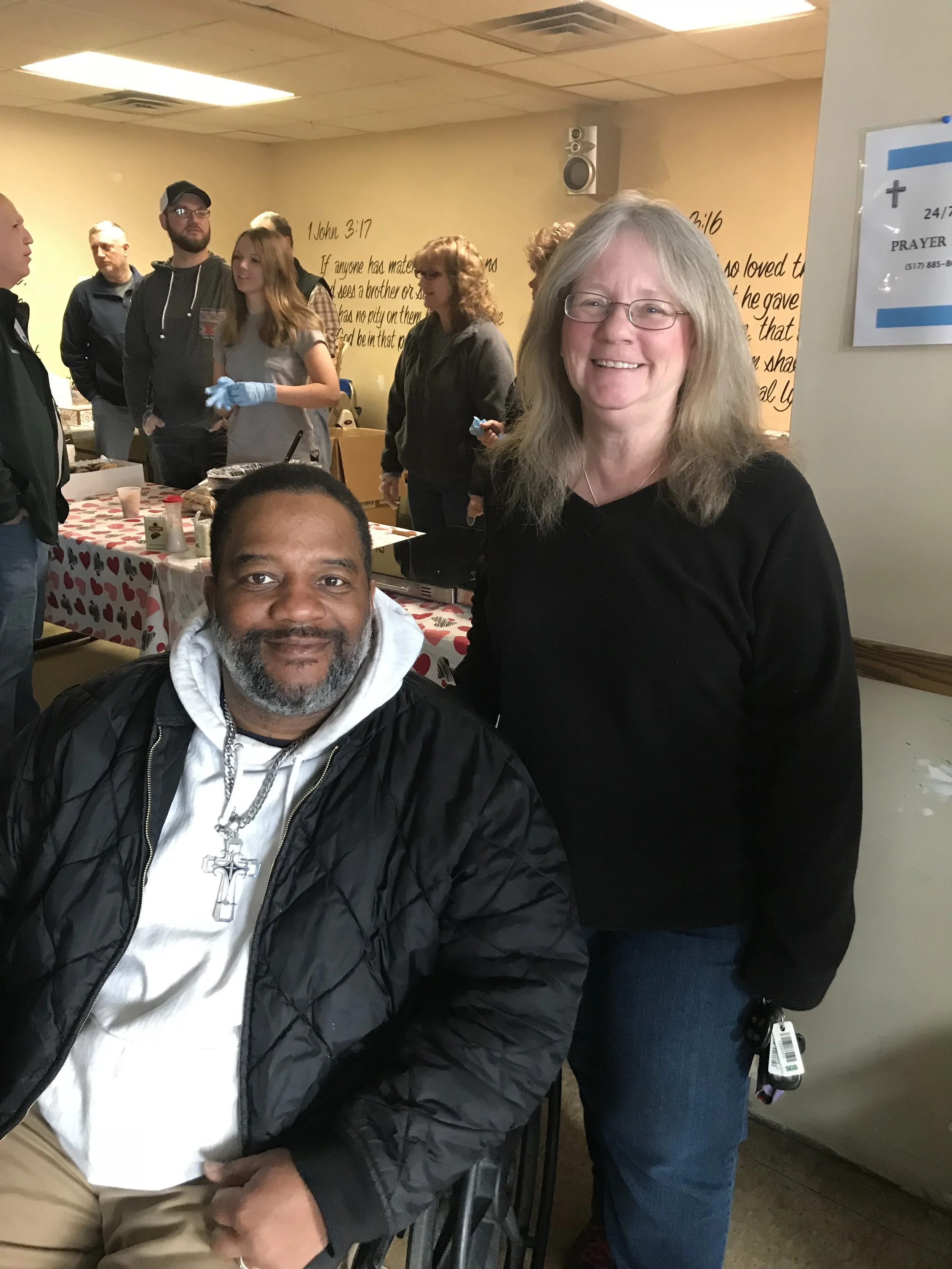 A group of people, including a man sitting in a wheelchair and a woman standing next to him, smiling at the camera. In the background, several people are gathered around a table with a heart-themed tablecloth at a community or church event.