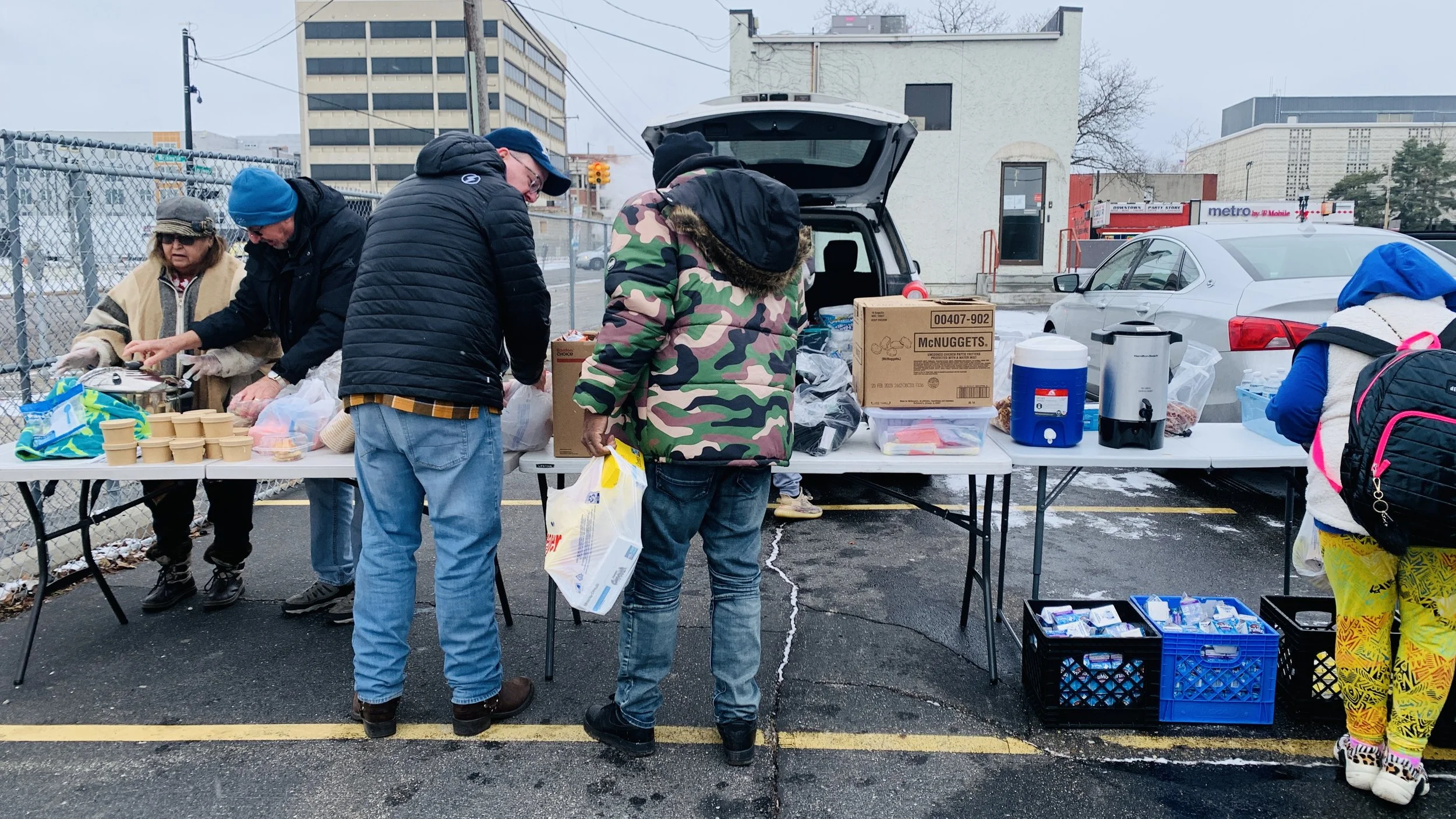 People standing at a table with supplies, in a parking lot with buildings and cars in the background, some wearing winter clothing.