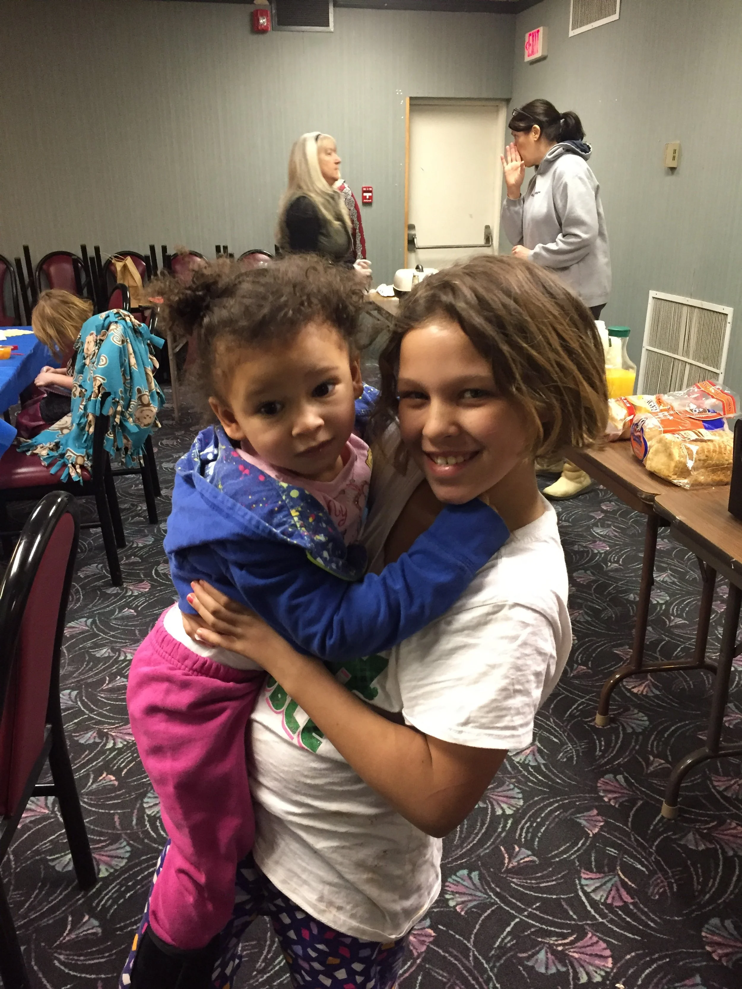 A young girl with short wavy hair holding a smaller girl with curly hair in a blue jacket, inside a room with tables and chairs, and other people in the background.