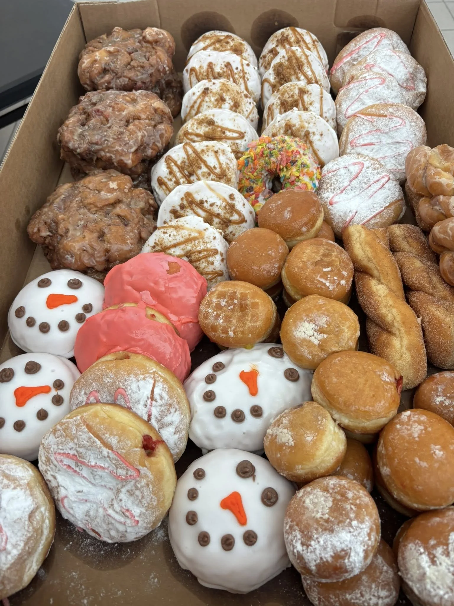 A box of assorted Christmas-themed donuts, including snowman faces with white icing, orange noses, and chocolate eyes, sprinkled and glazed varieties, and other decorated donuts with sprinkles and caramel drizzle.
