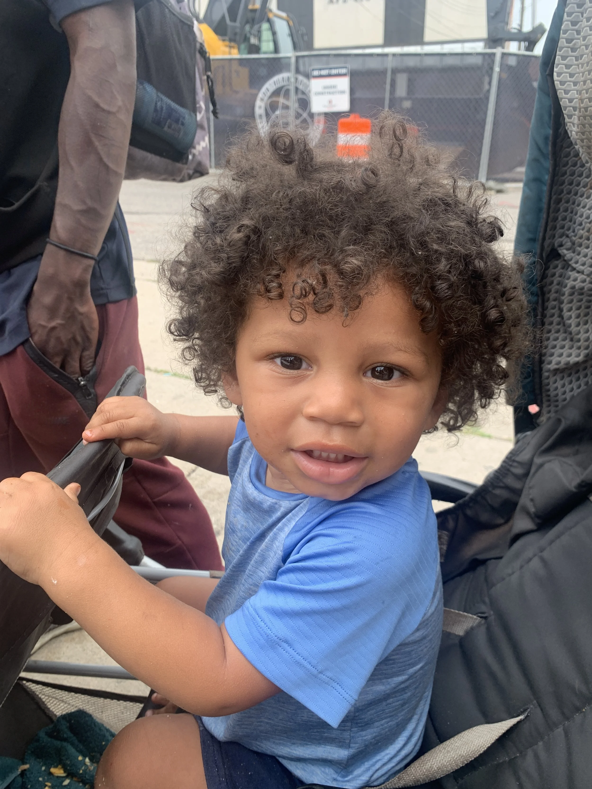 A young child with curly hair sitting in a stroller, looking up at the camera with a curious expression, wearing a blue shirt, surrounded by adults.