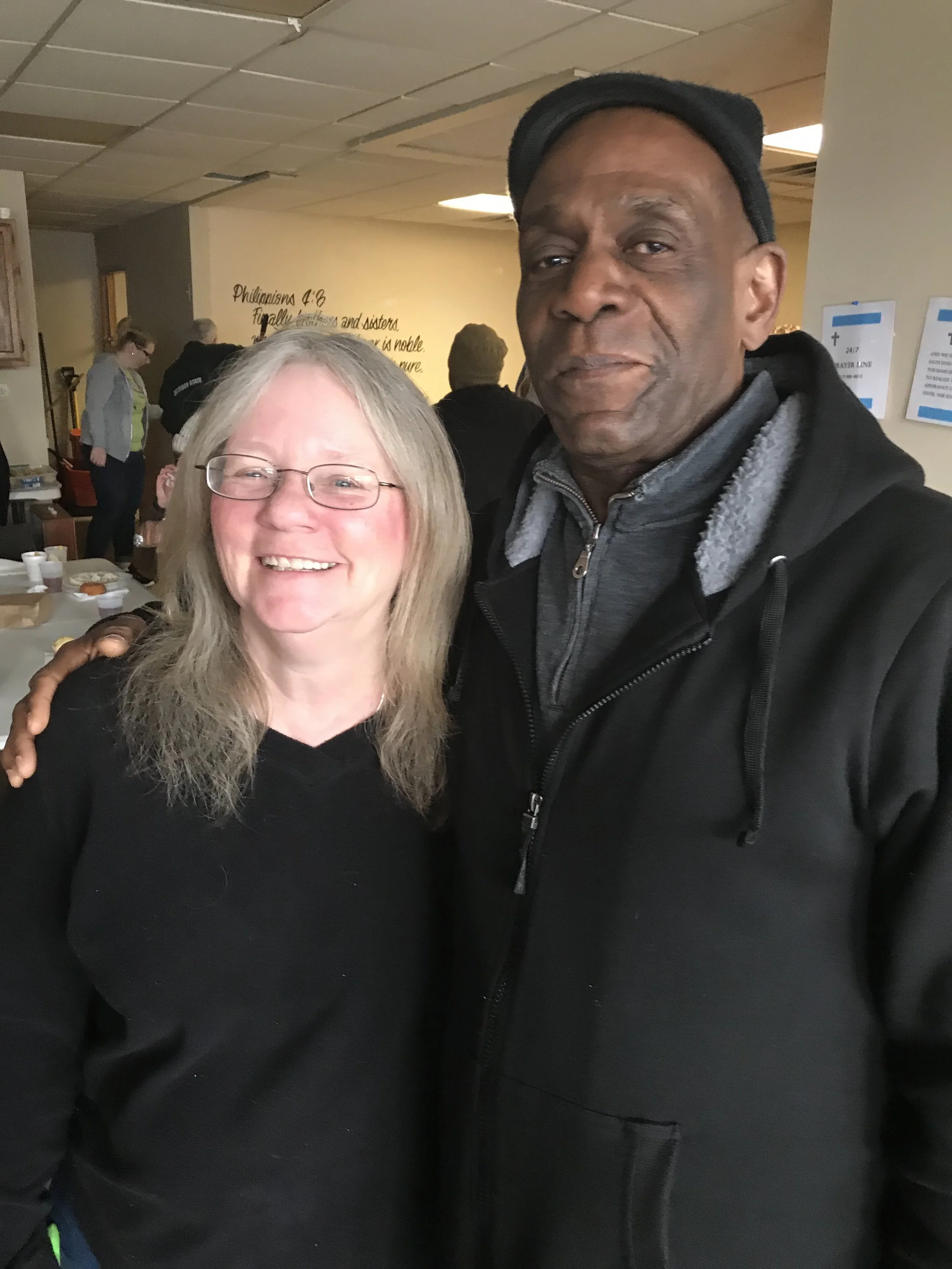 A smiling woman with glasses and long blonde hair standing next to a serious-looking man with a dark complexion, wearing a black jacket and cap. They are indoors at a gathering or event.