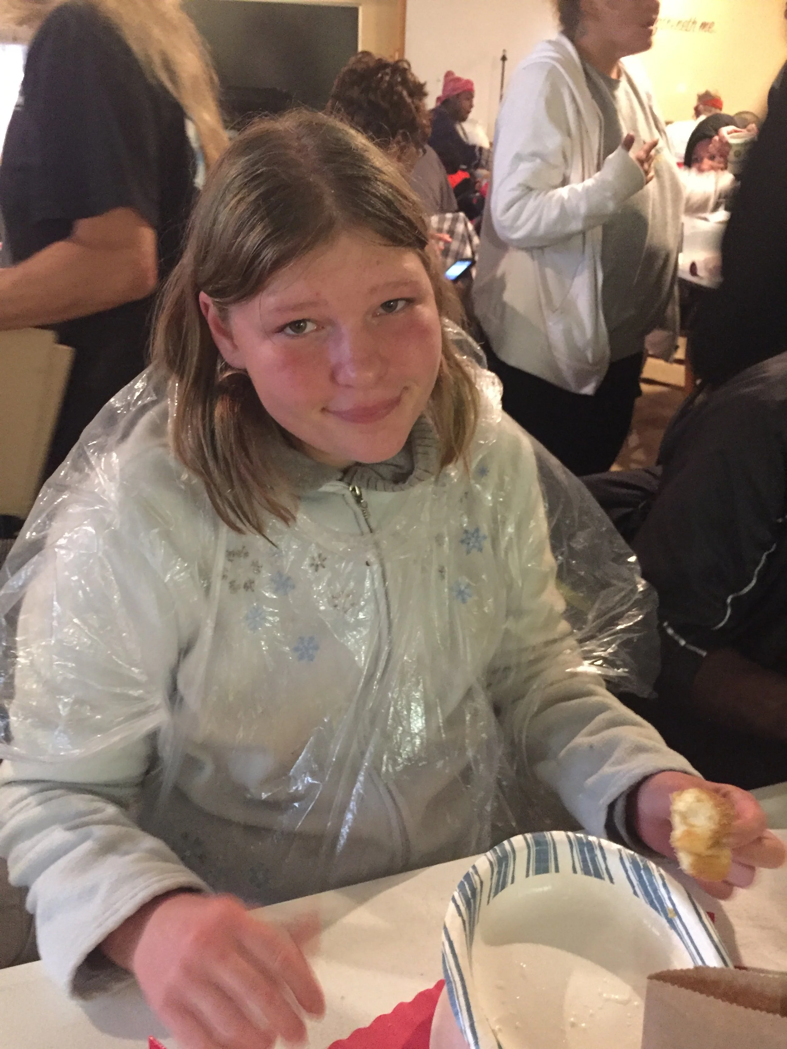 A young girl with light brown hair and a slight smile, wearing a white jacket with snowflake patterns and a clear plastic poncho, sitting at a table holding a piece of bread or pastry, in a busy indoor setting with people in the background.