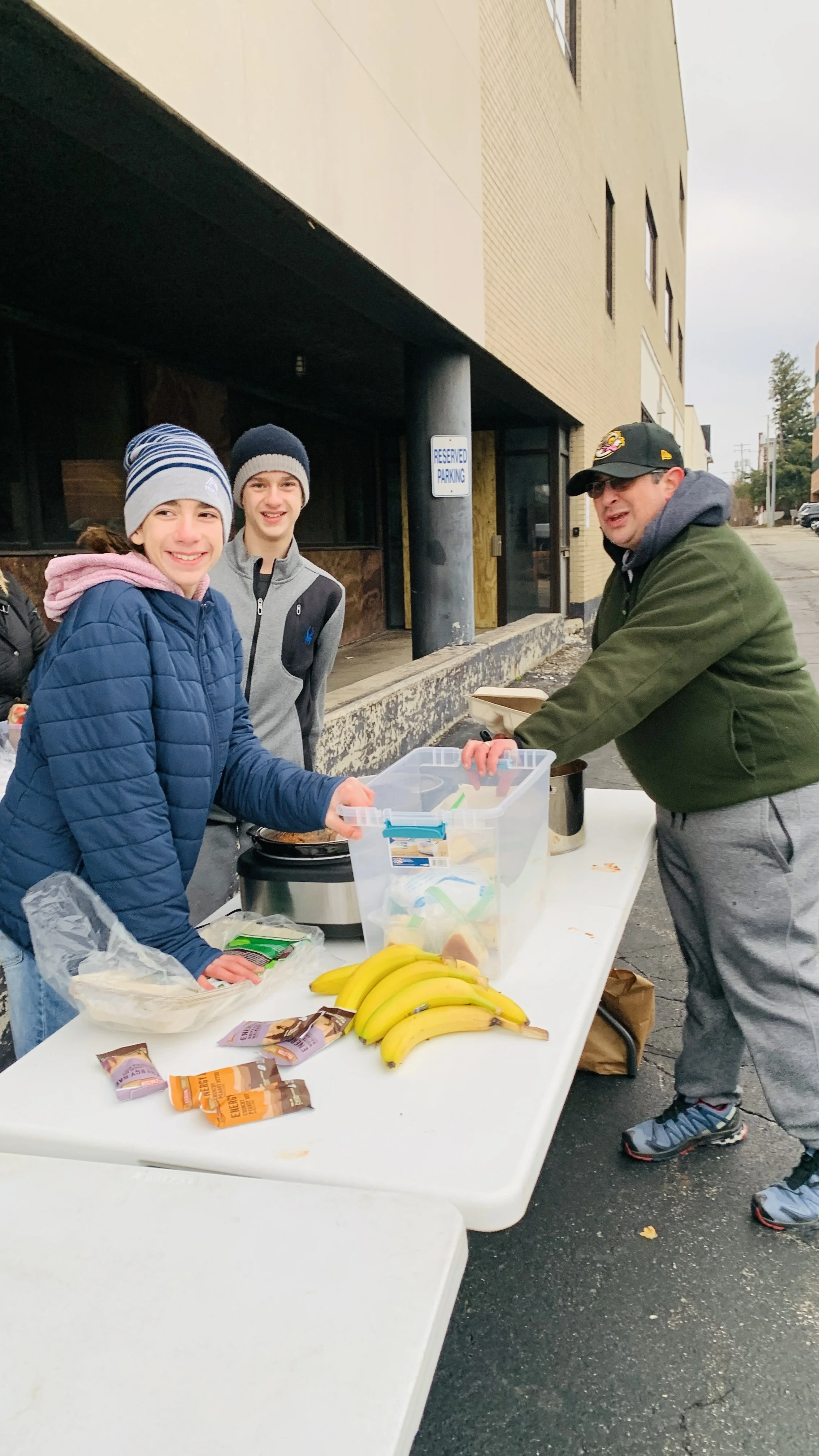 Two young boys and a man at an outdoor food table with bananas and snacks, smiling and engaged in serving food.