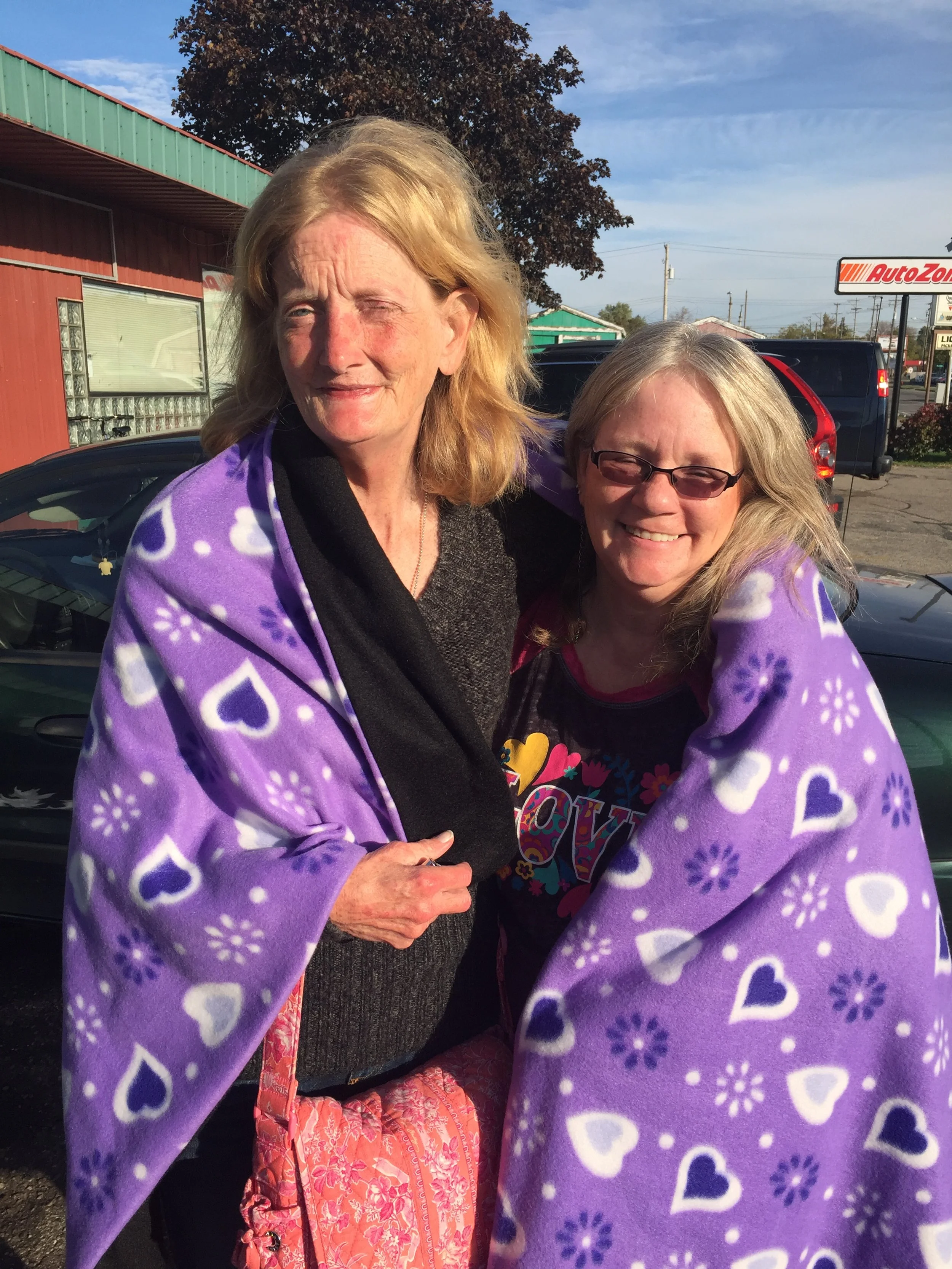 Two women smiling and embracing outdoors, wrapped in purple blankets with white hearts and floral patterns, standing in front of cars and a storefront under clear skies.