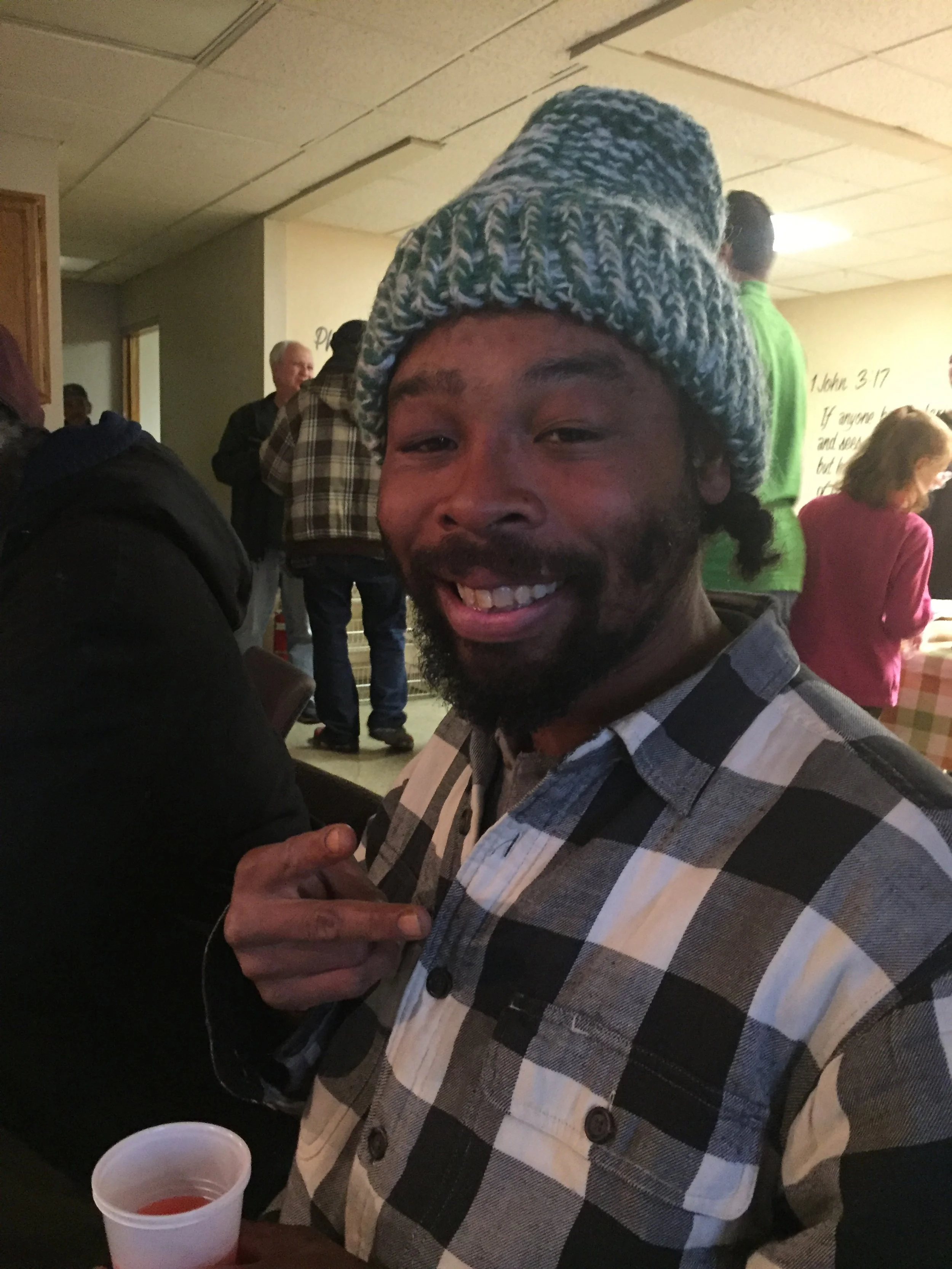 A man with a beard and a love of wearing a knitted gray, white, and blue beanie, smiling at the camera, holding a cup of red beverage, in a room with other people and a whiteboard background.