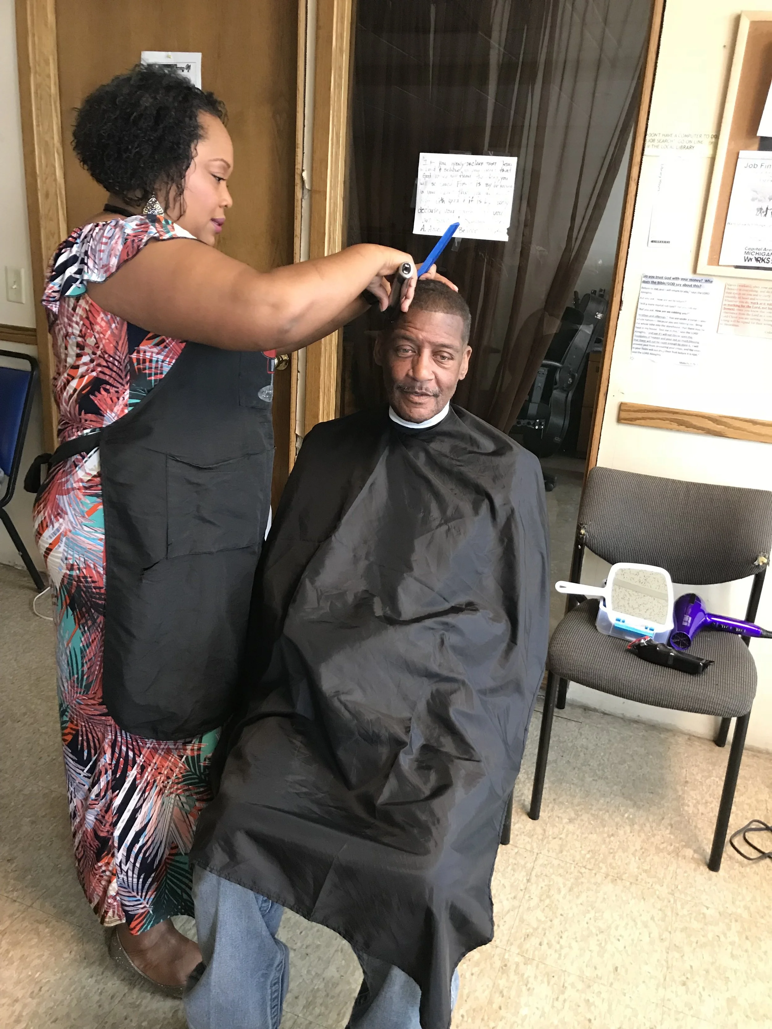 A woman getting a haircut from a barber in a barber shop. The man is seated in a chair with a black barber cape. The woman is standing and holding scissors and a comb. Behind the man, there is a chair with a purple hairdryer and a mirror on the wall.